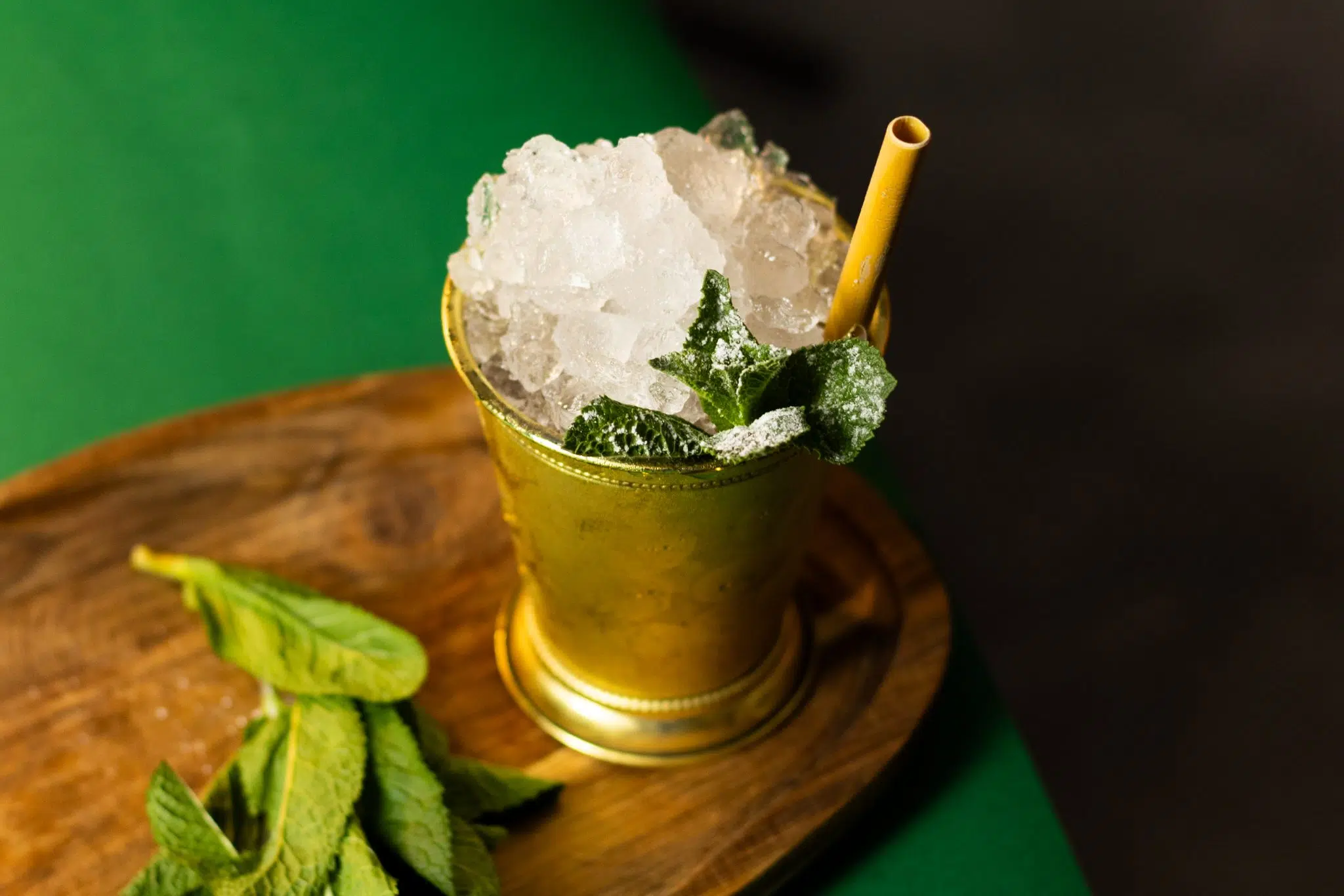 A high angle shot of a Brandy Julep cocktail in a julep cup on a wooden board placed on a green surface with some mint leaves on the side