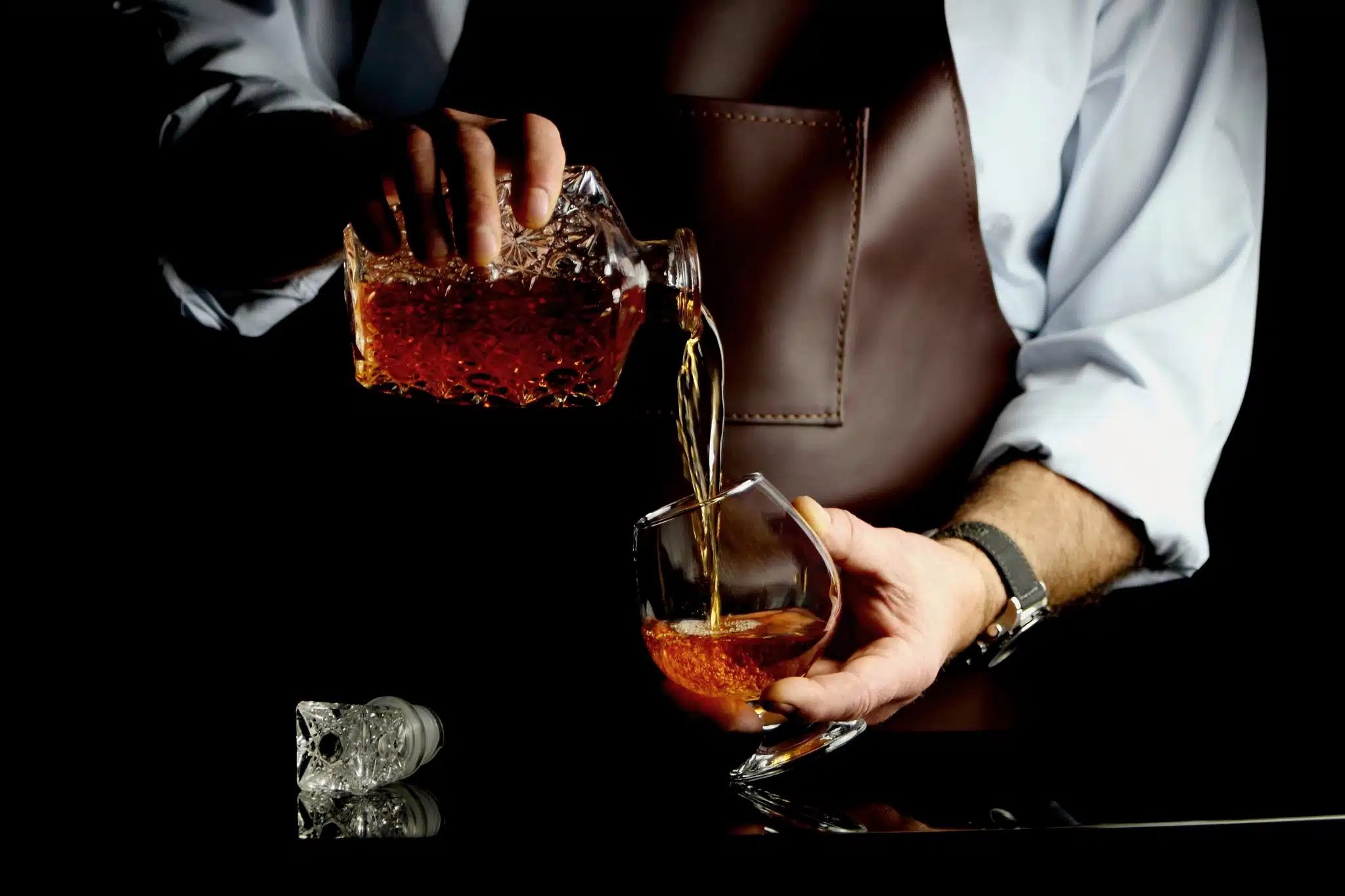 A person in a dress shirt pours amber-colored brandy from a crystal decanter into a glass against a dark background.