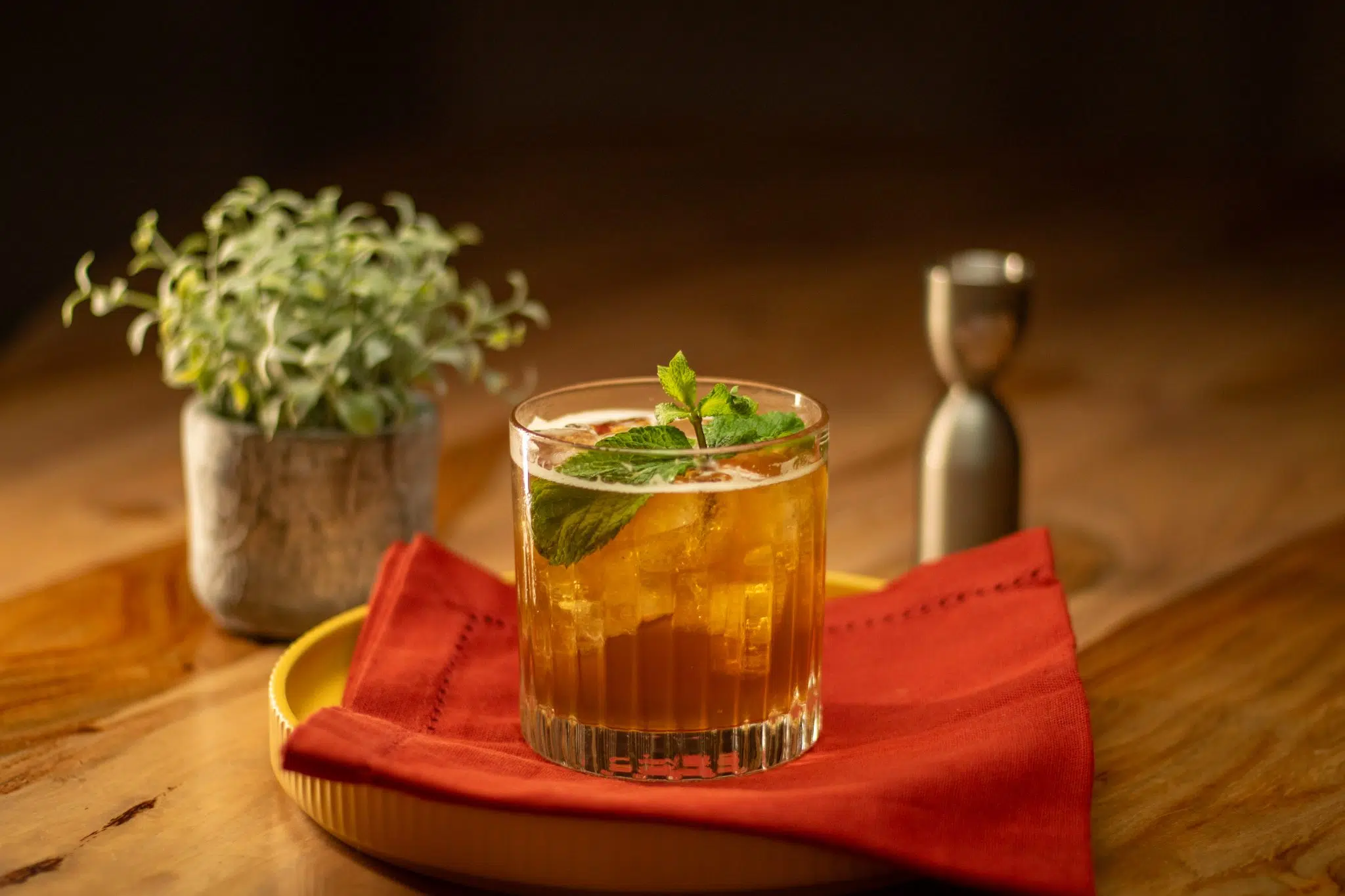 A side shot of a Brandy Smash cocktail in an old fashioned glass on a red cloth placed on a yellow tray and a wooden surface surrounded by a jigger and a plant in a pot