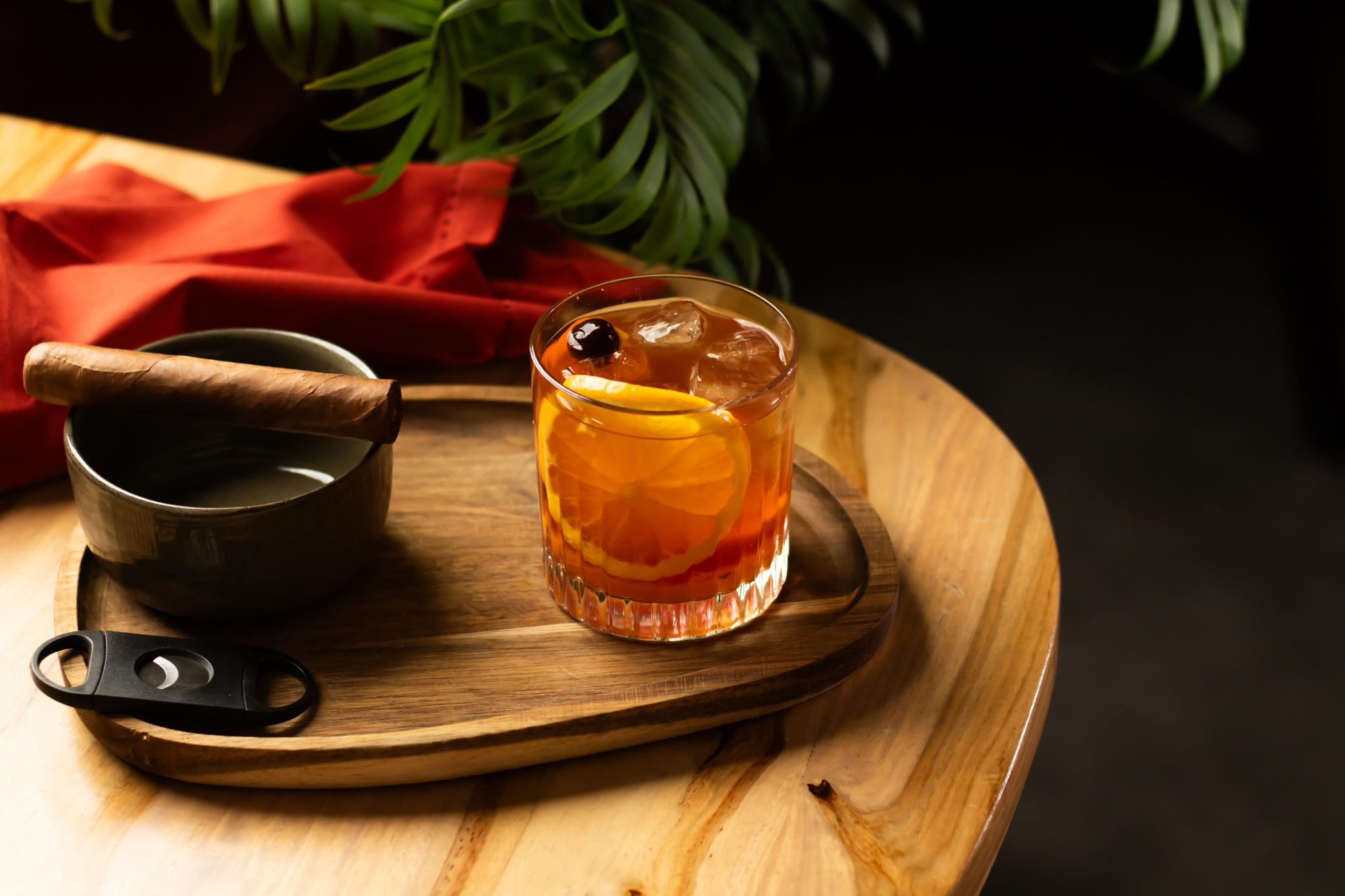 A side shot of a Brandy Wisconsin Old Fashioned cocktail in an old fashioned glass on a wooden board placed on a wooden table surrounded by a brown bowl, a cigar, a red cloth, and a cigar cutter.