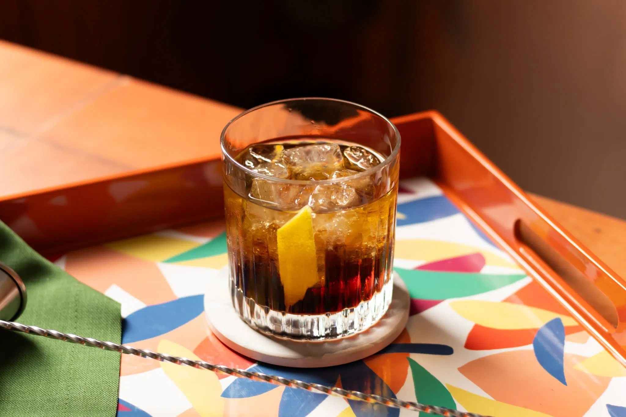 A side shot of a Brave Bull cocktail in an old fashioned glass on a white coaster on a multicolor tray surrounded by a bar spoon and a green cloth