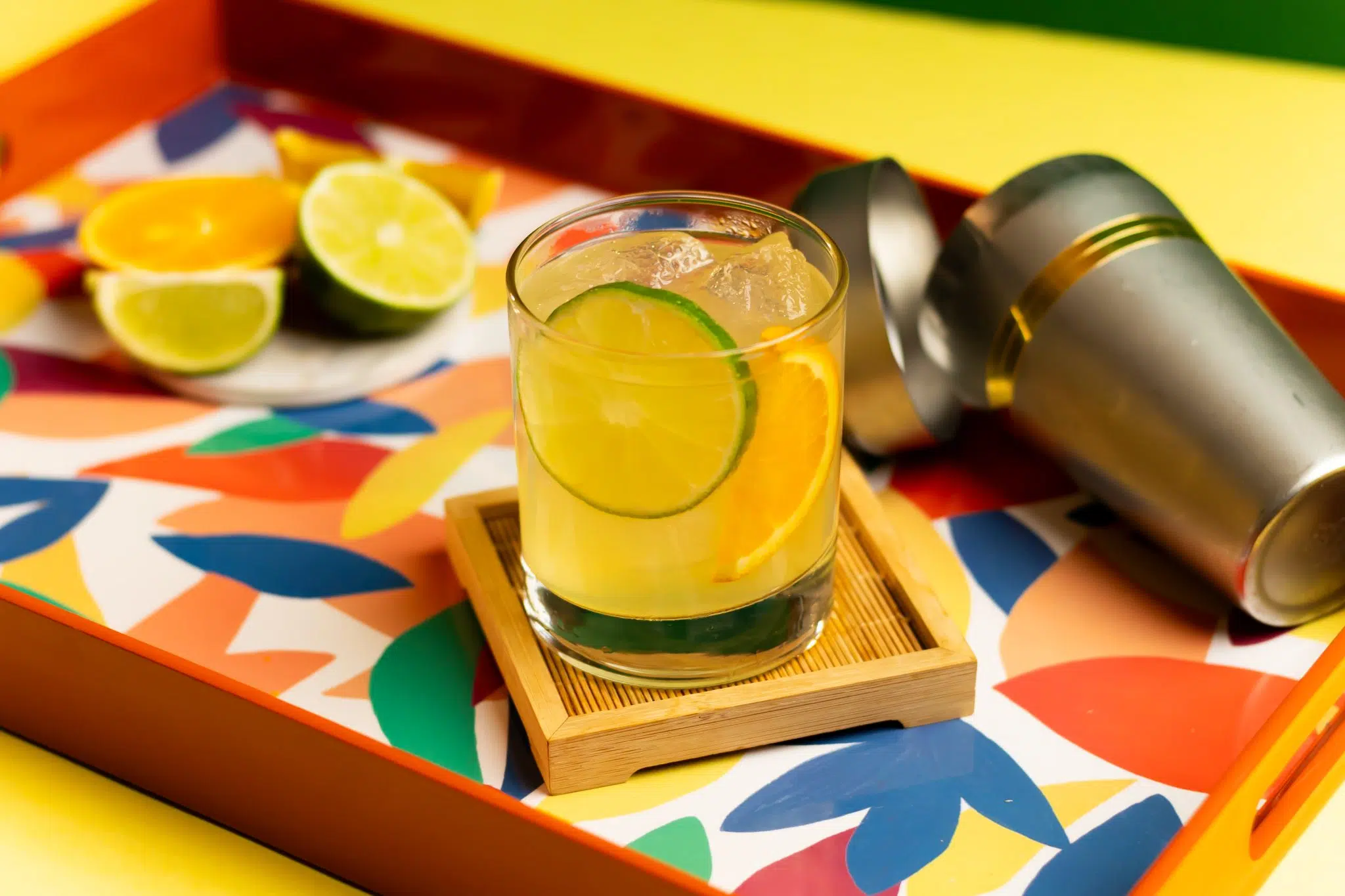 A side shot of a Brazil 66 cocktail in an old fashioned glass on a wooden coaster placed on a multicolor tray surrounded by a shaker, two lime pieces and an orange slice