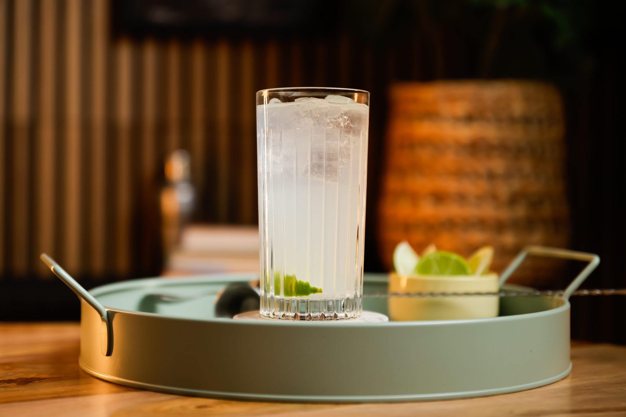 A side shot of a Brazilian Buck cocktail in a highball glass on a turquoise tray surrounded by a bowl with lime pieces, a jigger and a bar spoon