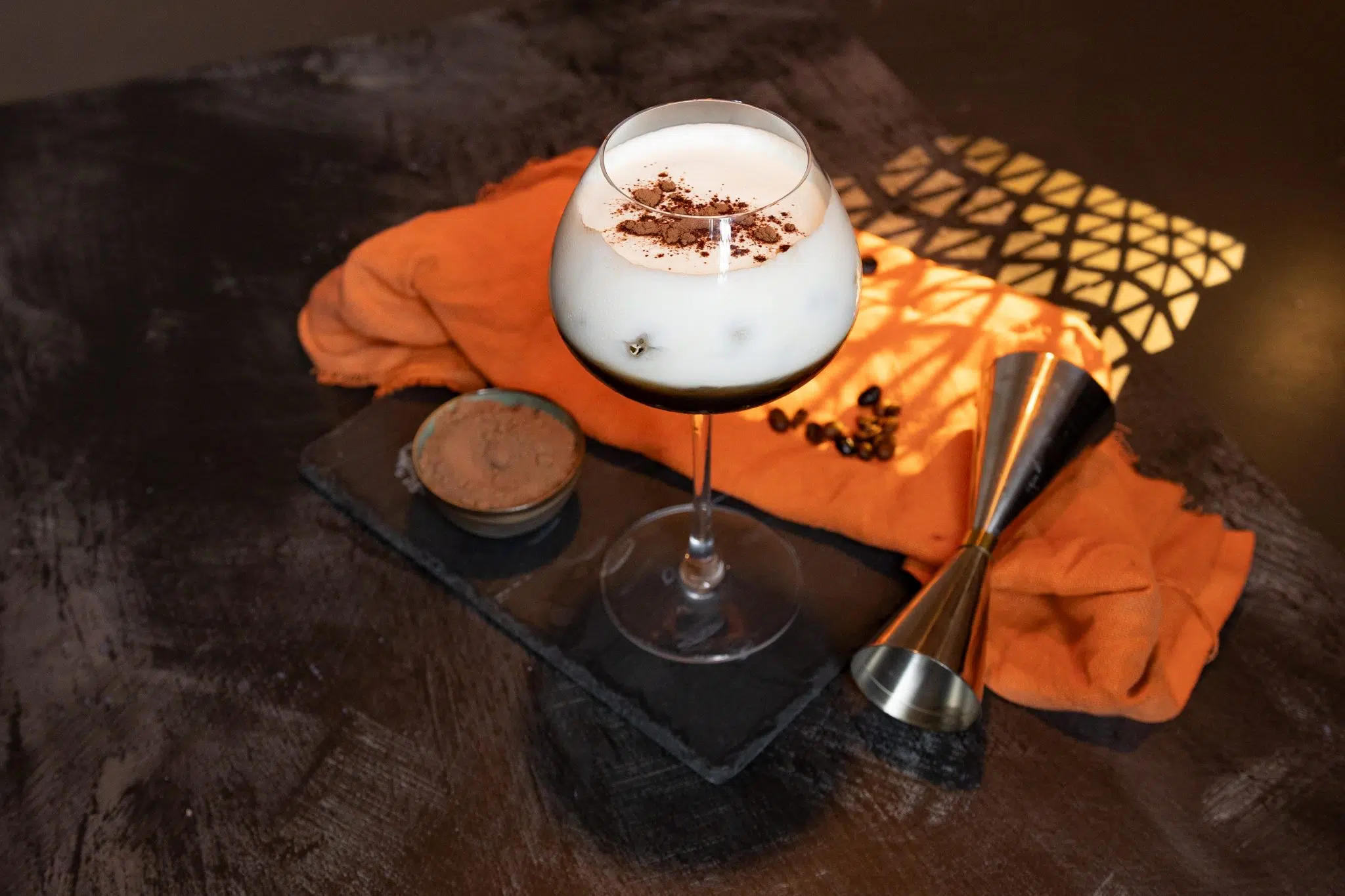 A side shot of a Brazilian Coffee cocktail in a coupe glass on a black stone board place on a black surface surrounded by an orange cloth, a jigger, a bowl with cocoa powder and coffee beans