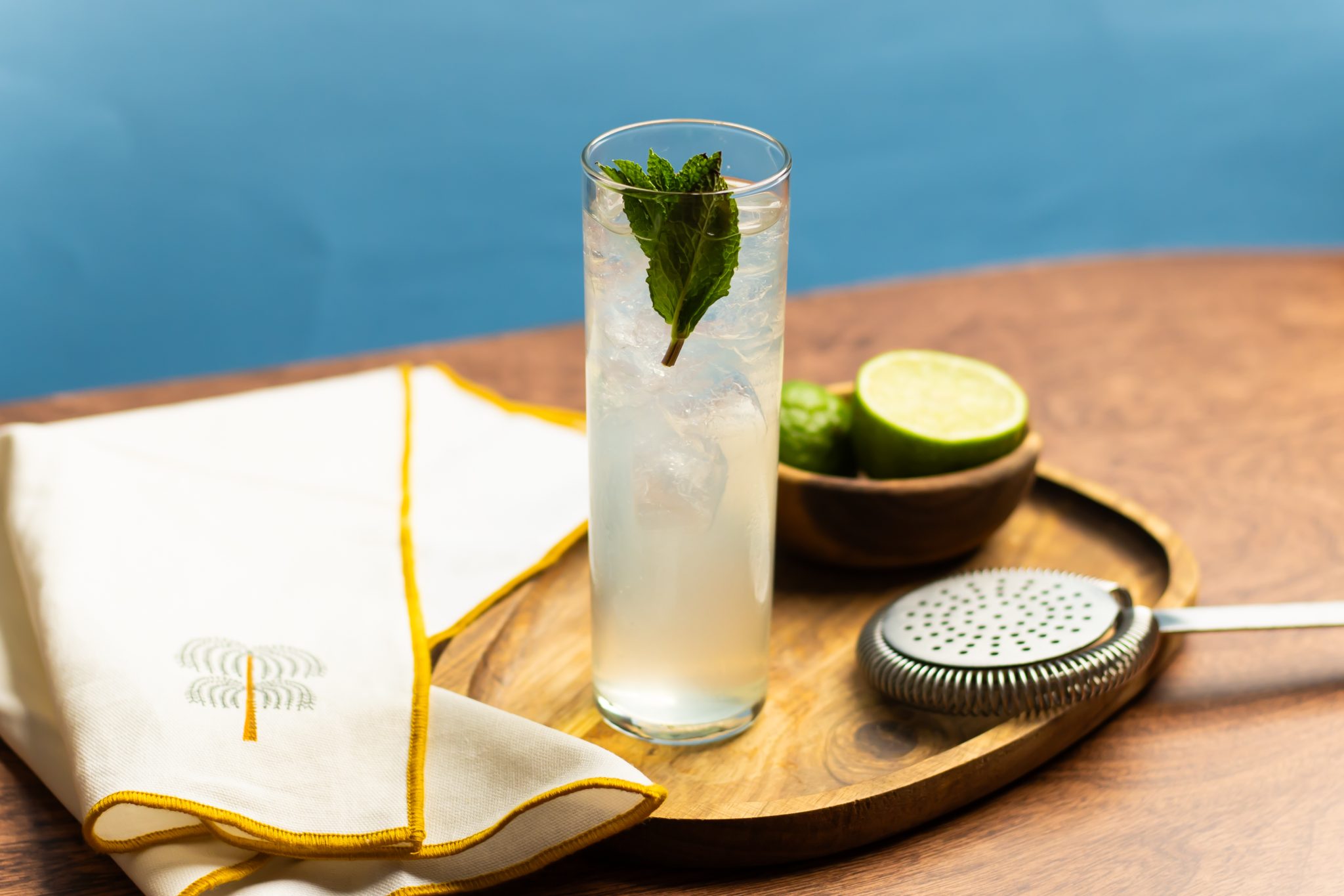 A side shot of a Brazilian Mule cocktail in a highball glass on a wooden board surrounded by a strainer, a white cloth and a bowl with lime wedges