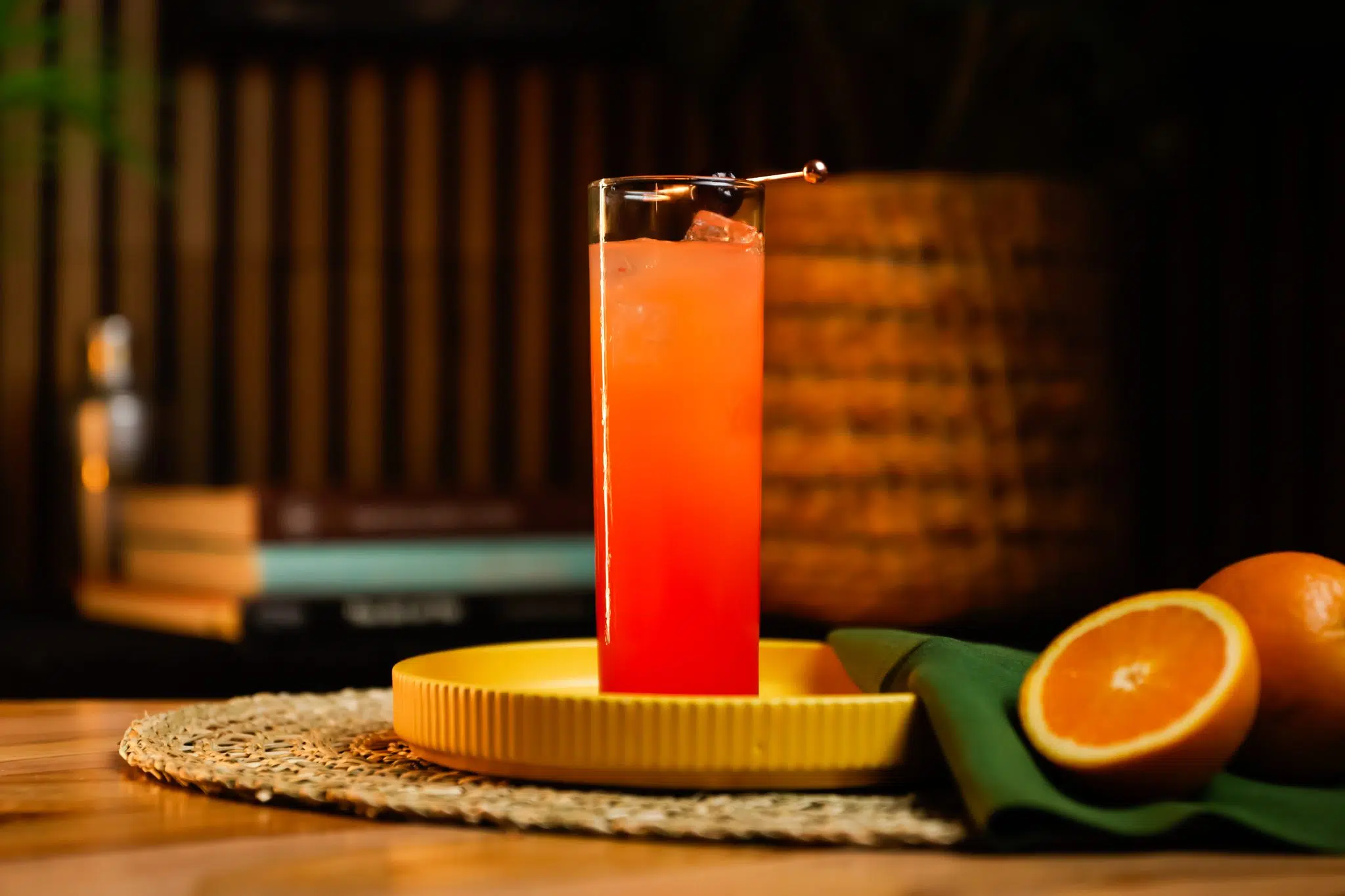 A side shot of a Brazilian Sunrise cocktail in a highball glass on a yellow tray placed on a straw placemat on top of a wooden table, with two oranges and a green cloth on the side