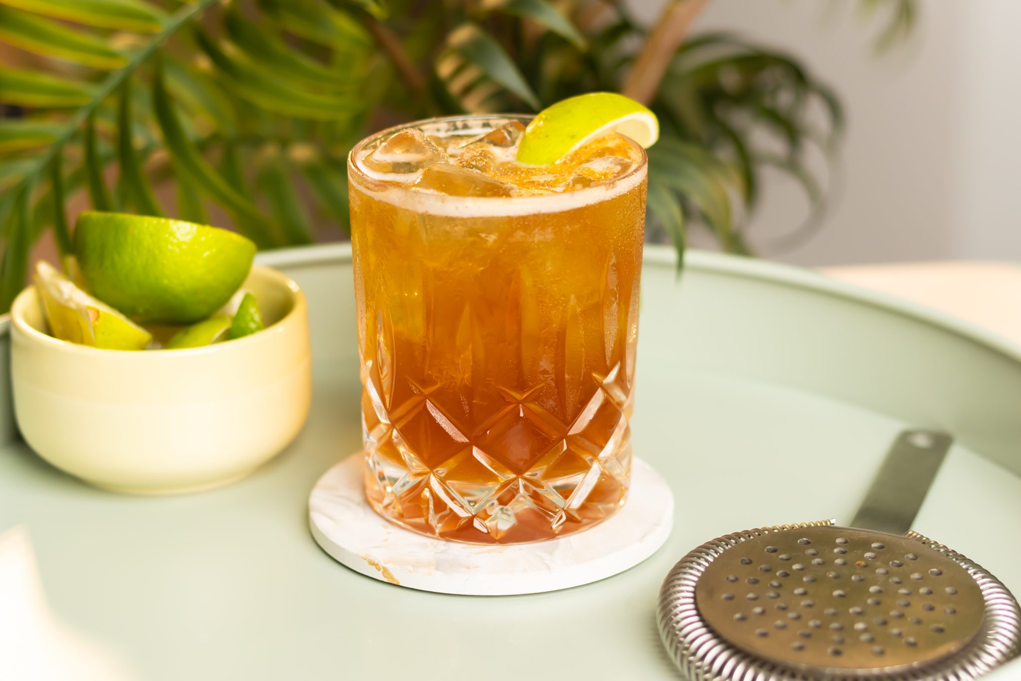 A side shot of a Bumbo drink in an old fashioned glass on a white coaster on a light green tray surrounded by a strainer, and a white bowl with lime pieces.