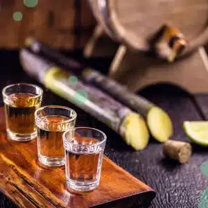 Three diferent shot glasses with cachaça on a dark brown wooden tray with a lime wedge, a cork, two pieces of sugar cane and a barrel behind.