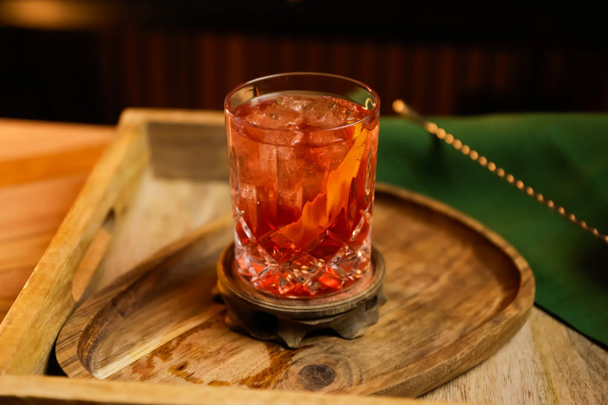 A side shot of a Cachaça Old Fashioned cocktail in an old fashioned glass on a wooden coaster and tray surrounded by a straw, a bar spoon, and a green cloth