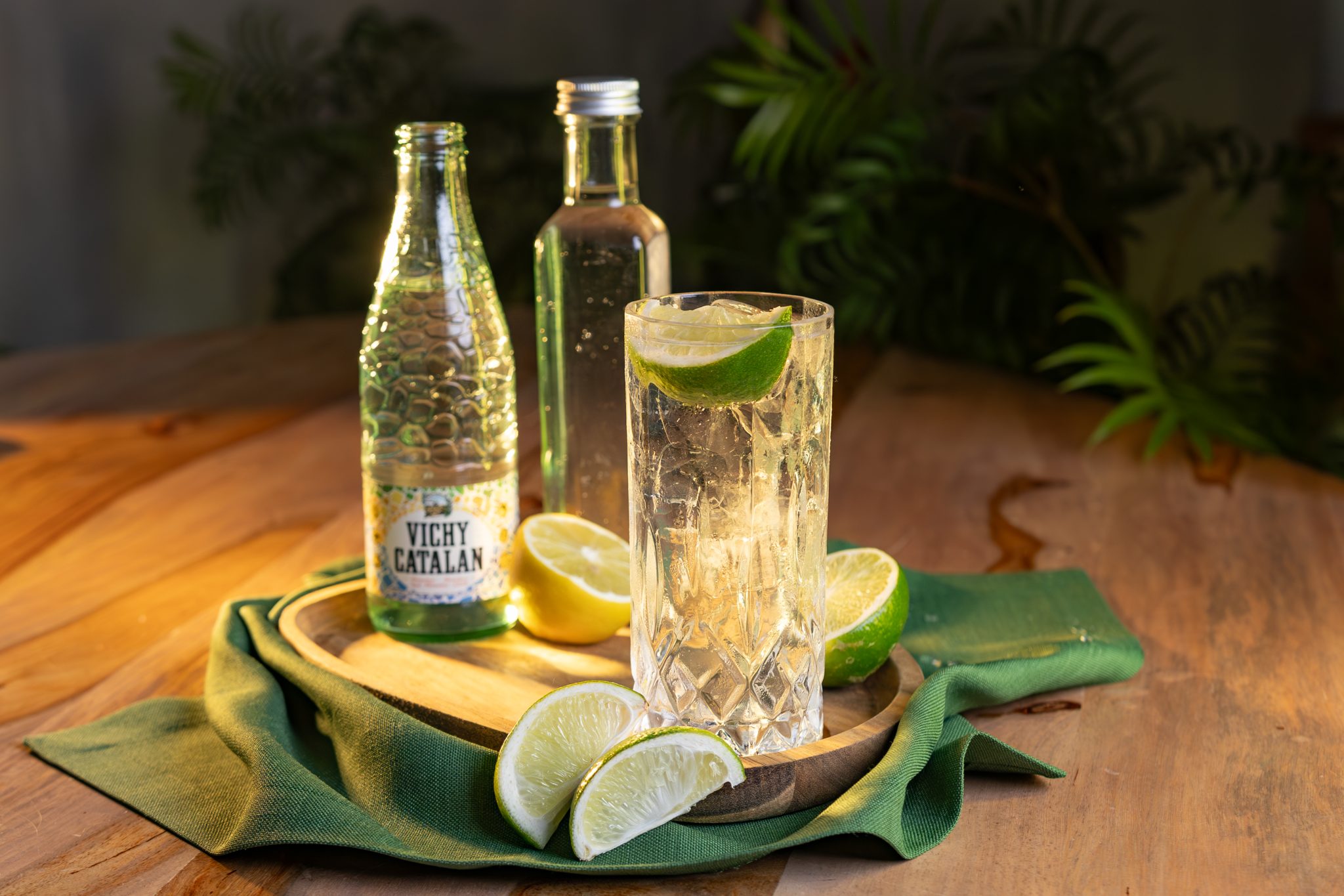 A side shot of a Cachaça and Soda cocktail in a highball glass on a wooden tray placed on a wooden table surrounded by soda, half a lemon, two lime slices, half a lime, and a green cloth