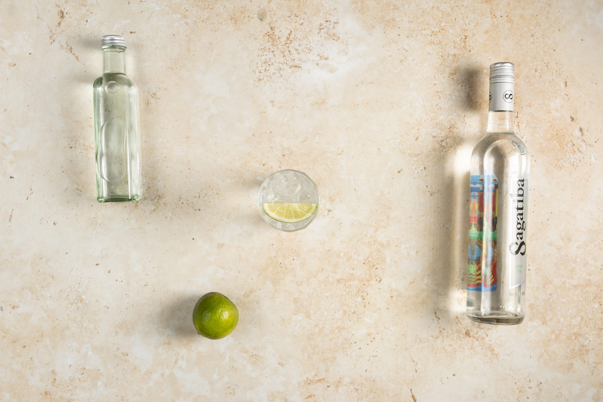 A Cachaça and Soda cocktail, shot from above, in a highball glass on a beige surface surrounded by Cachaça, soda and lime