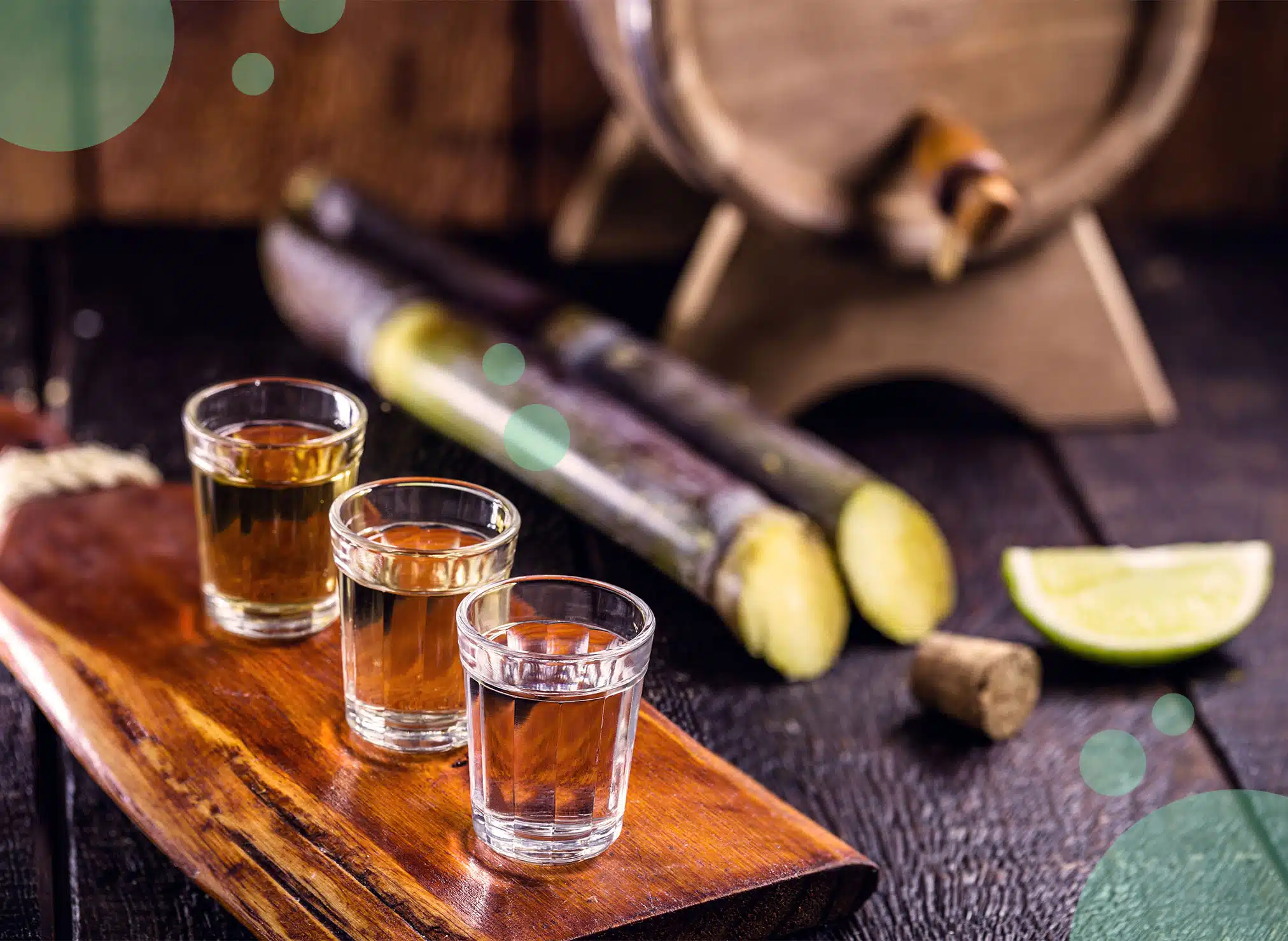 Three diferent shot glasses with cachaça on a dark brown wooden tray with a lime wedge, a cork, two pieces of sugar cane and a barrel behind.