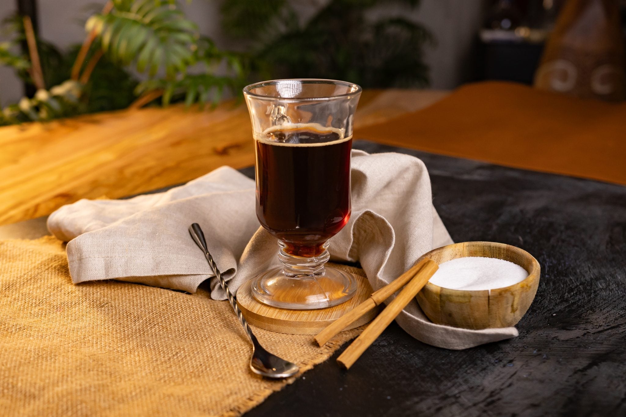 A side shot of a Café Quemado cocktail in a mug on a wooden coaster placed on a wooden surface surrounded by two cinnamon sticks, a bar spoon, a white cloth and a wooden bowl with sugar