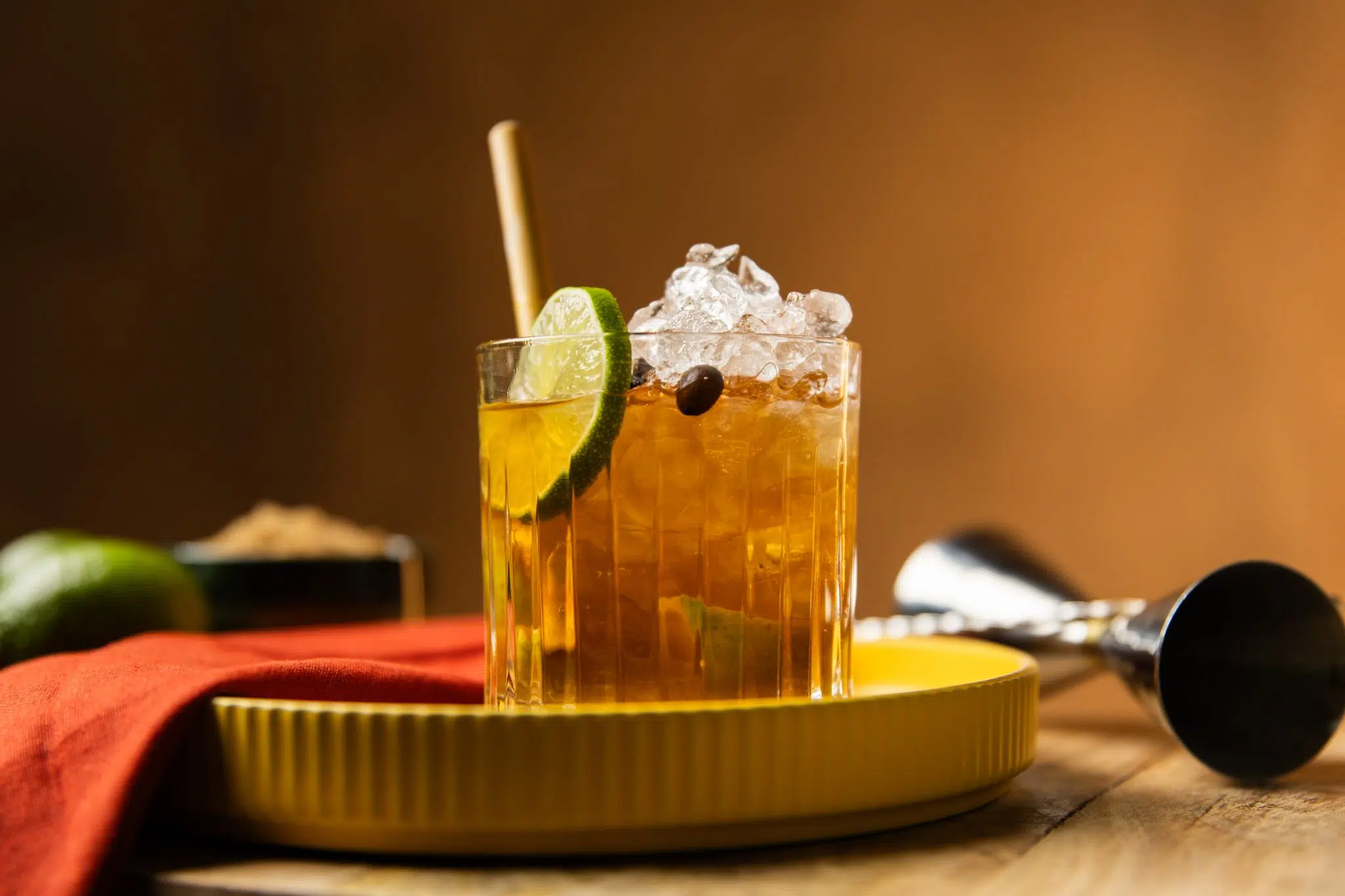 A side shot of a Caipirinha Coffee cocktail in an old fashioned glass on a yellow tray surrounded by a red cloth, a jigger, a lime and a bowl with demerara sugar