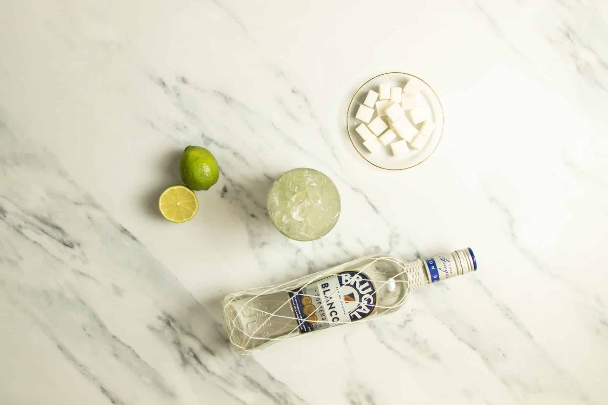 Caipirissima cocktail on a white marmol table with a white rum bottle, sugar cubes and limes around.