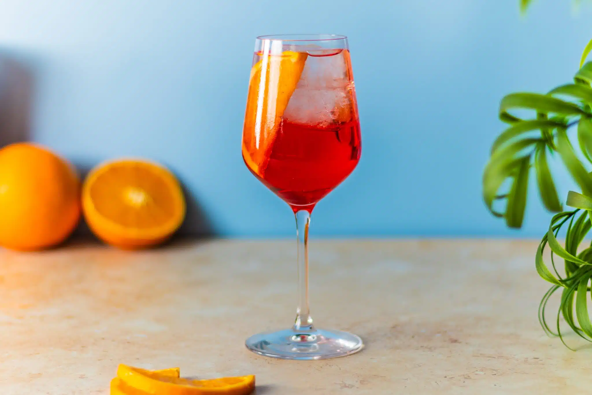 A side shot of a Campari Spritz cocktail in a wine glass on a beige table with orange slices in front and a half orange behind.