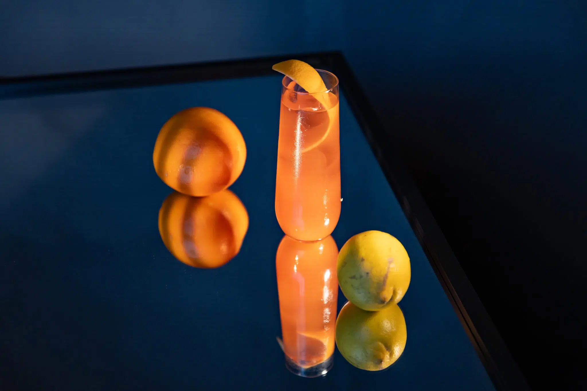 A side shot of a Campari and Grapefruit cocktail in a highball glass on a mirror surface surrounded by an orange and a lemon