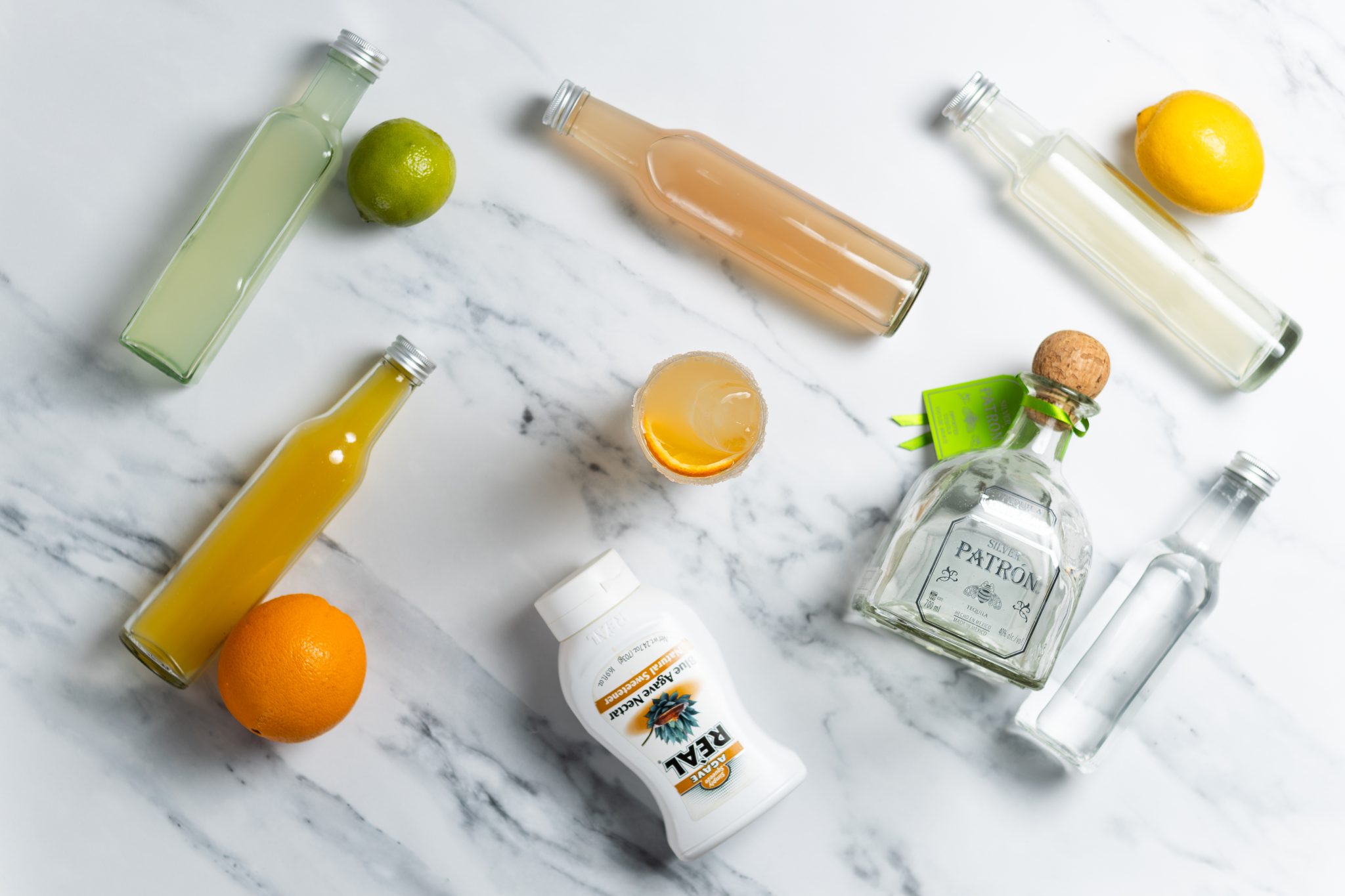 A Cantarito cocktail, shot from above, in a highball glass on a white marmol table surrounded by lemon juice, lime juice, grapefruit juice, orange juice, agave syrup, soda water, and Tequila.