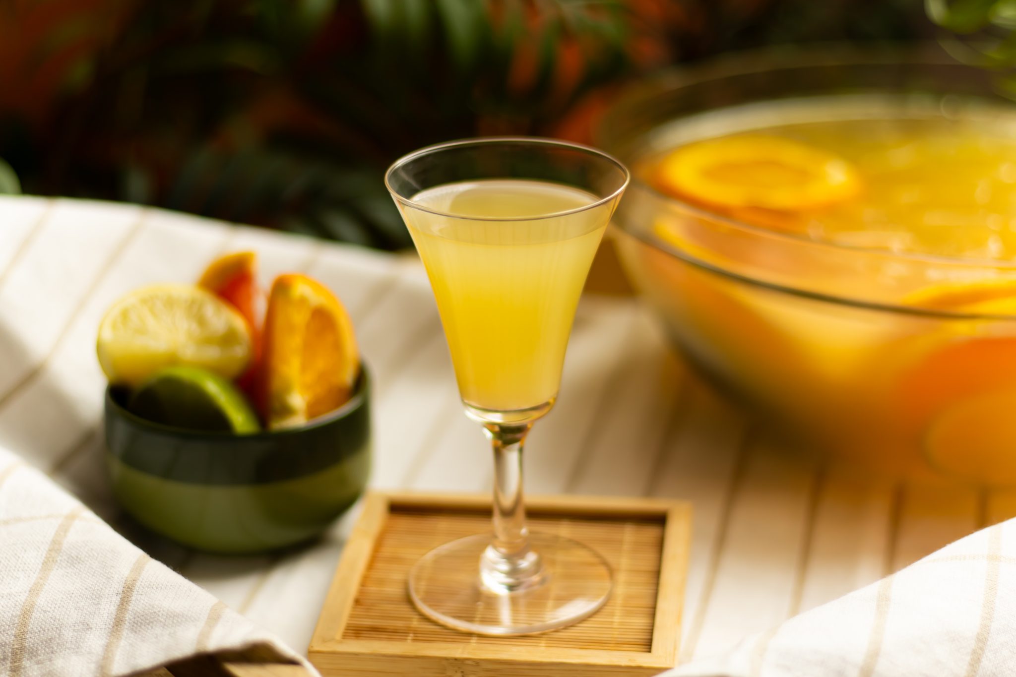 A side shot of a Cazuela drink in a highball glass on a wooden coaster placed on a white cloth with stripes surrounded by a punch with more Cazuela drink and a green bowl with lime, lemon, grapefruit, and orange wedges.