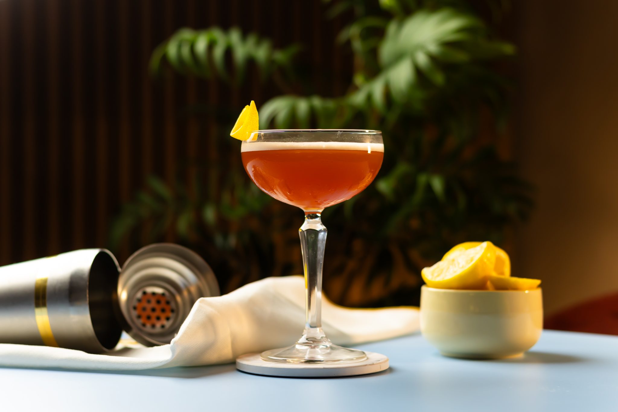 A side shot of a Champs-Elysees cocktail in a cocktail glass on a white coaster placed on a light blue table surrounded by a shaker, a white cloth and a white bowl with lemon pieces.