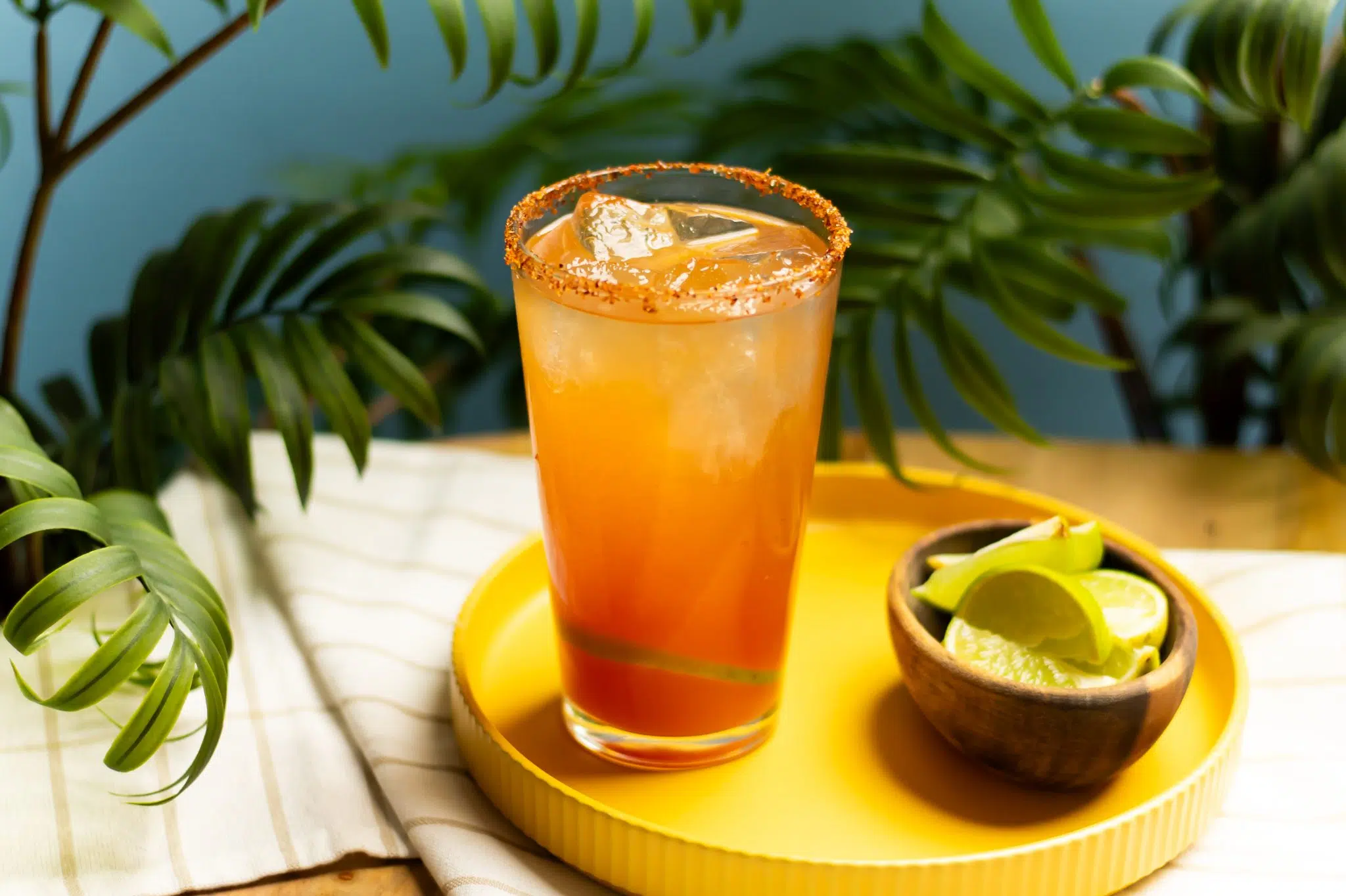 A side shot of a Chavela Drink in a pint glass on a yellow tray surrounded by a white cloth a brown bowl with lemon wedges, and plant leaves in front of a blue background