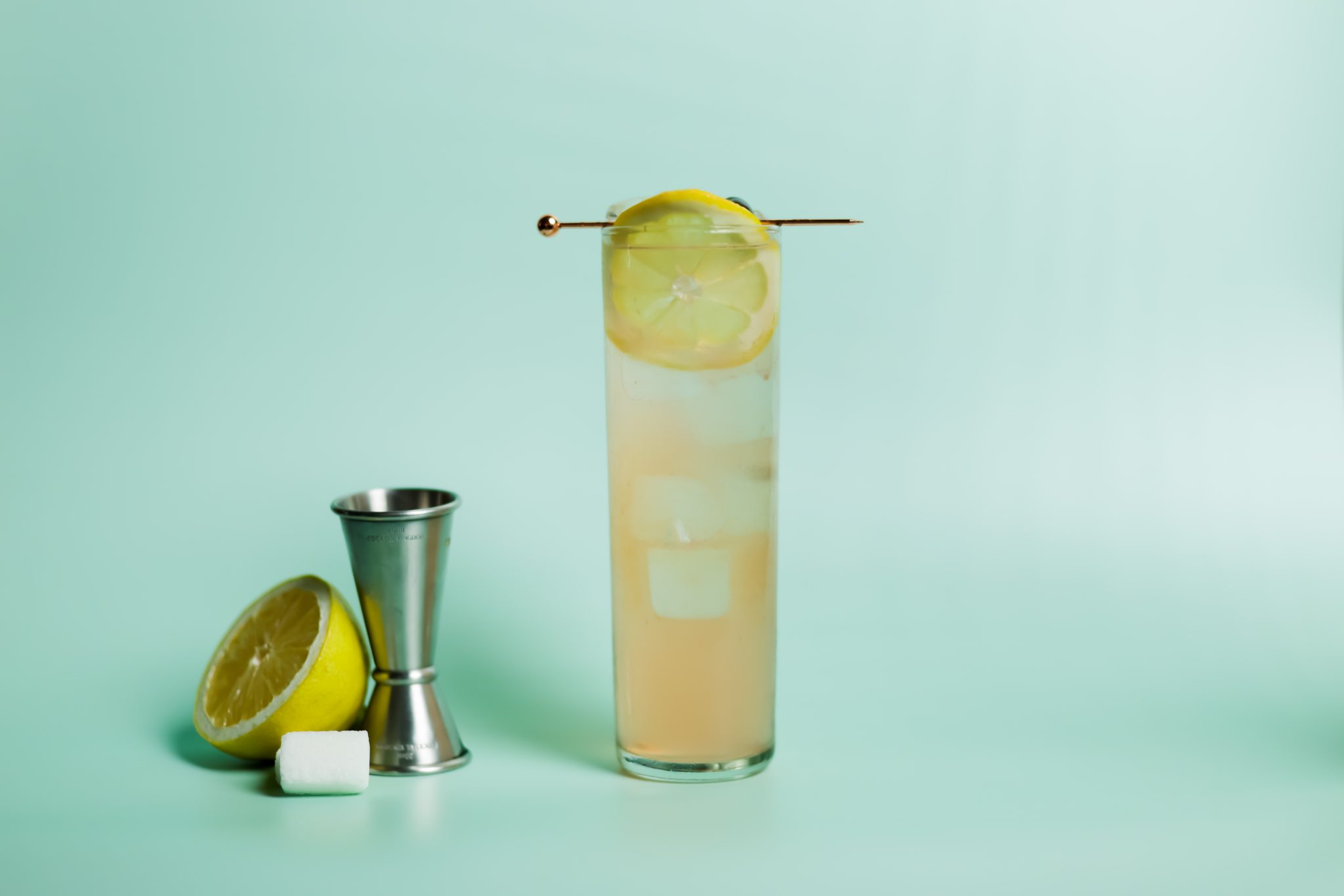 A side shot of a Cherry Collins cocktail in a highball glass on a turquoise background color with a jigger, half a lemon and two sugar cubes aside.