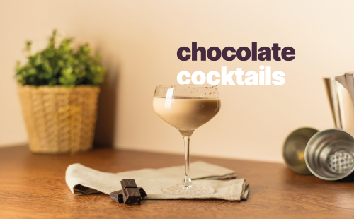 A side shot of a chocolate cocktail in a coupe glass on a wooden table surrounded by a shaker, some chocolate squares and a plant behind