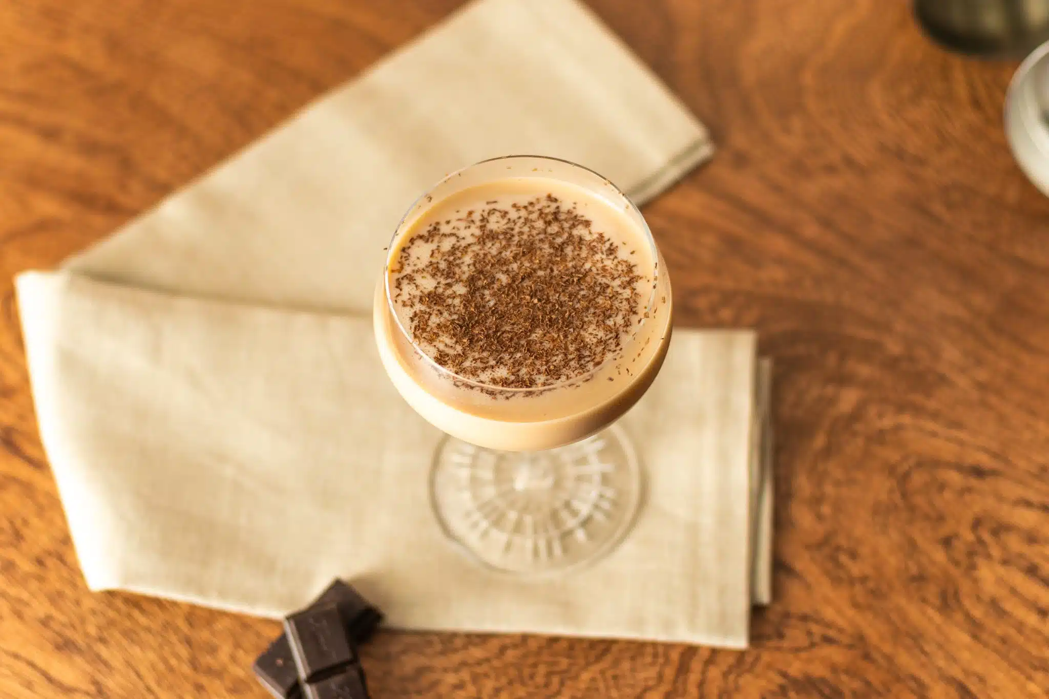 A Chocolate Martini cocktail, shot from above, in a martini glass on a beige cloth with chocolate squares on a side, placed on dark brown table.