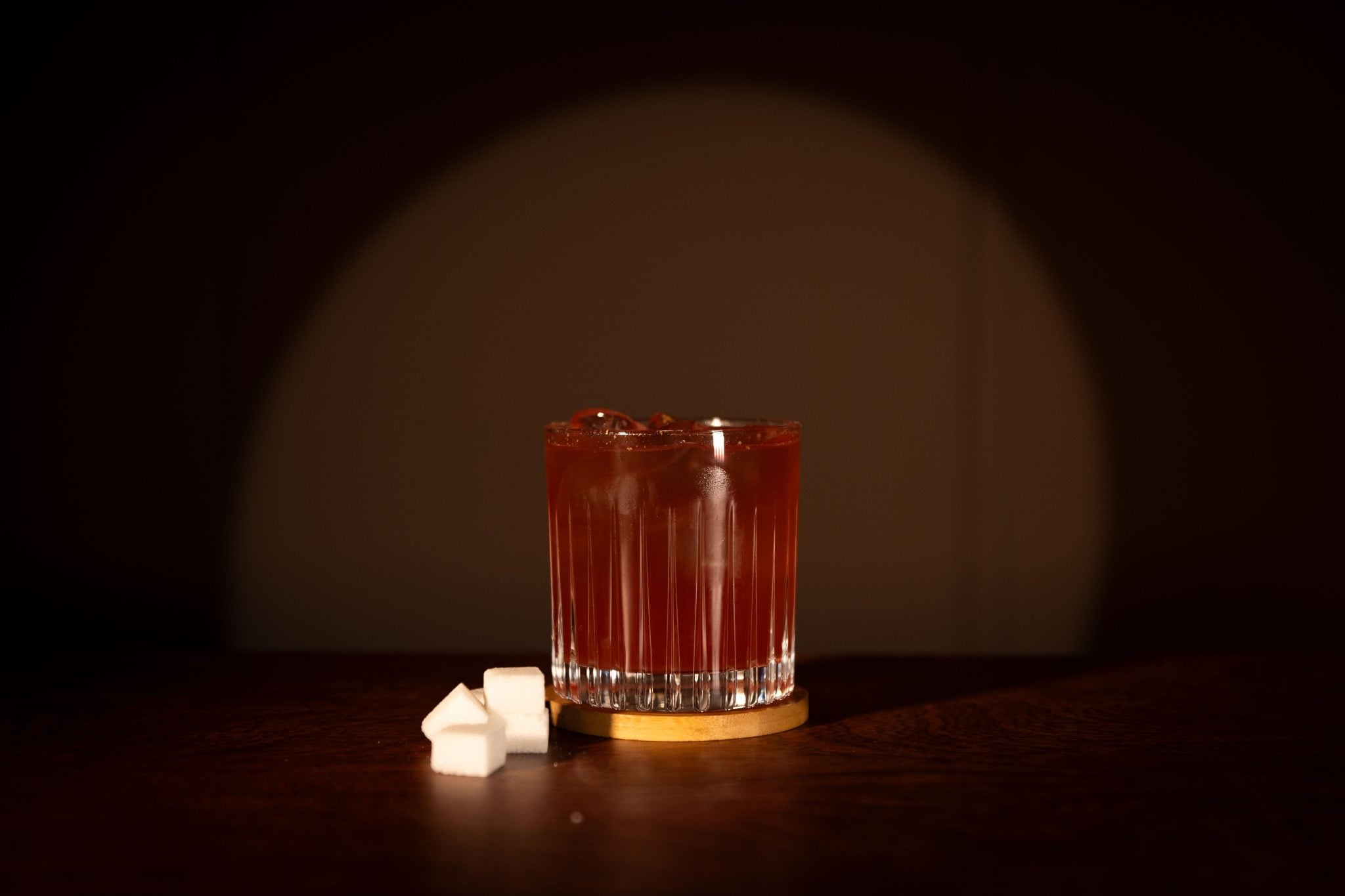 A side shot of a Christmas Punch cocktail in an old fashioned glass on a wooden coaster placed on a wooden surface with sugar cubes on the side.