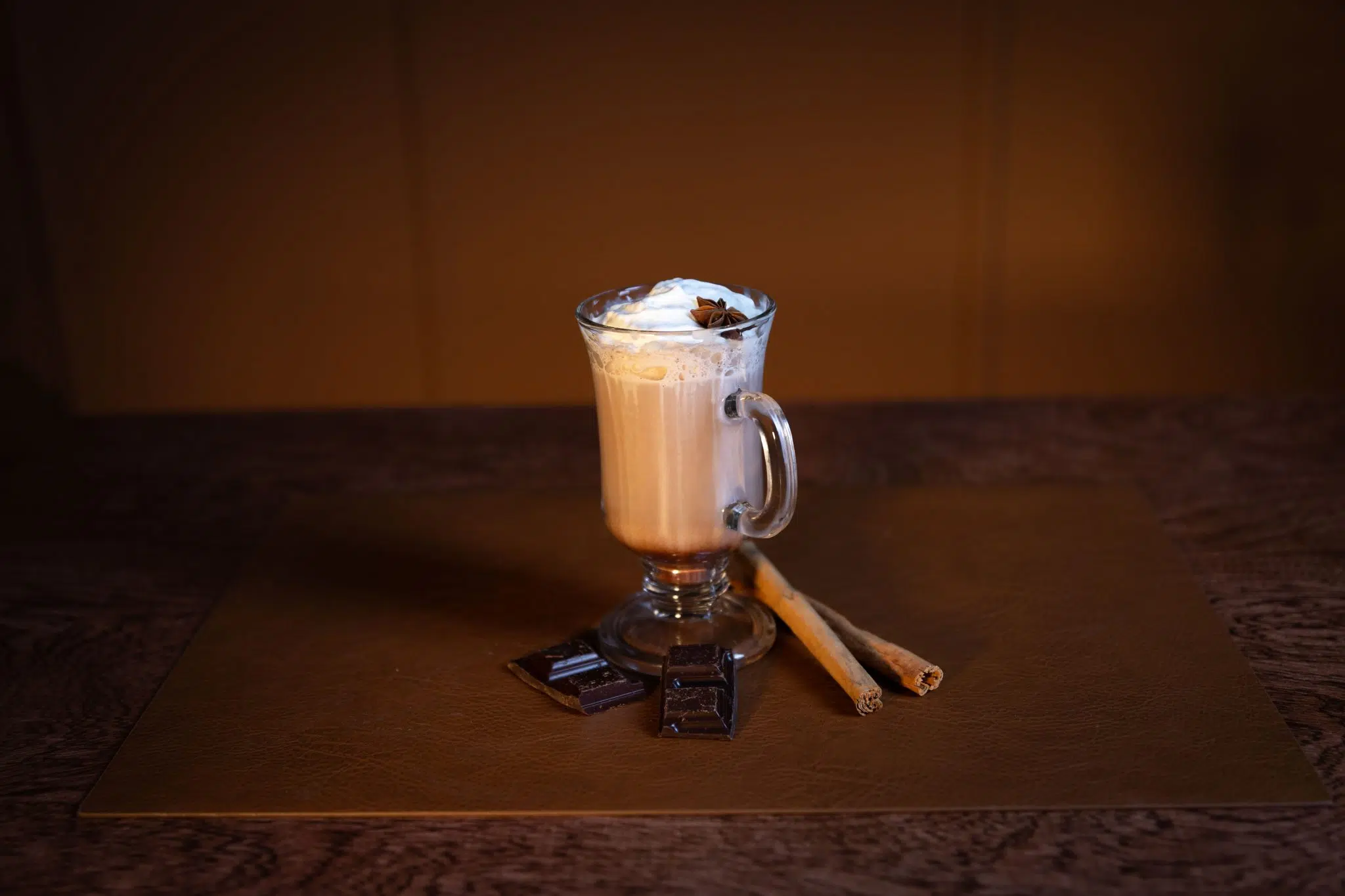 A side shot of a Cinnamon Aguardiente Hot Chocolate cocktail in a mug on a brown surface surrounded by two chocolate squares and two cinnamon sticks