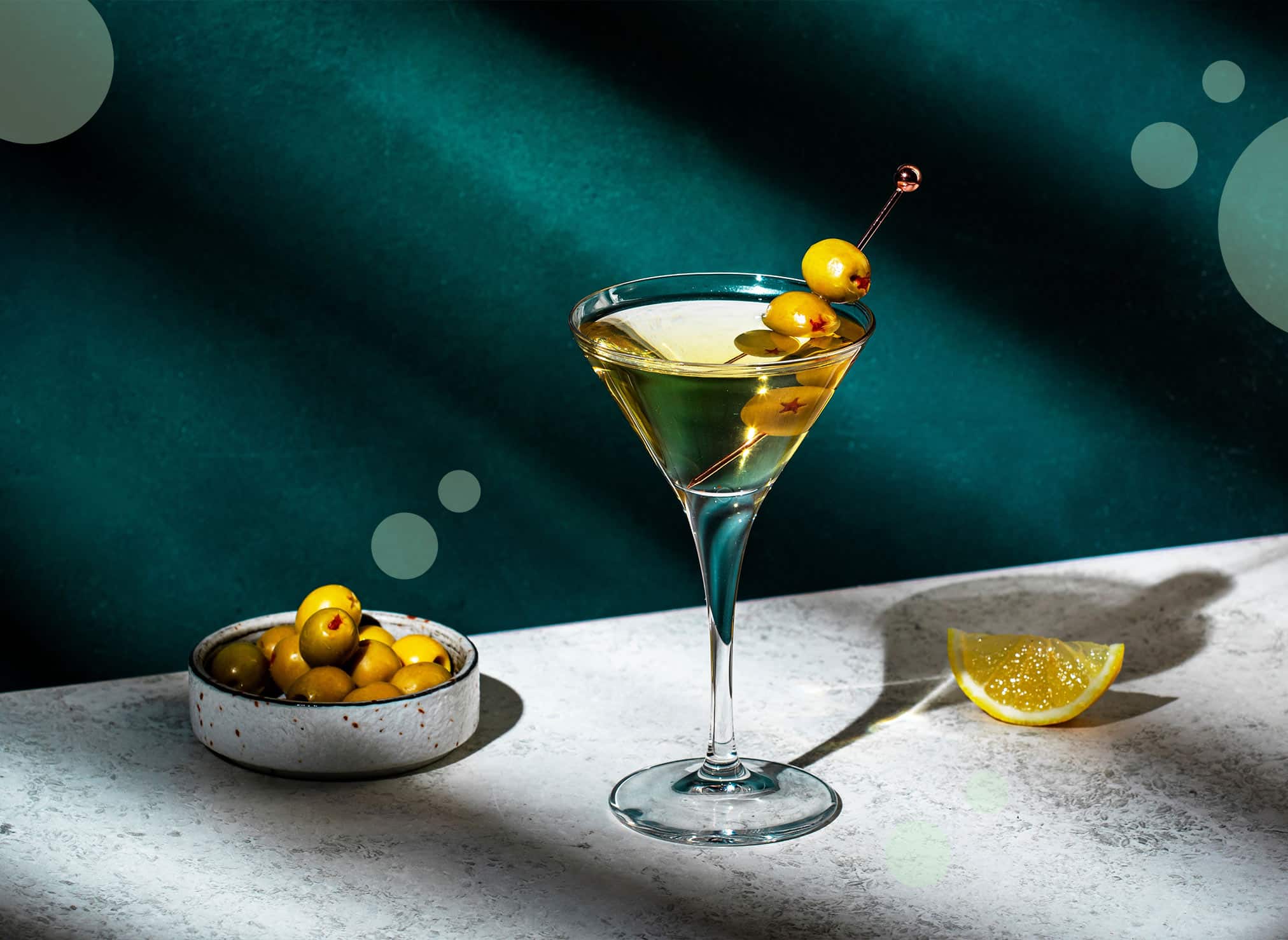 Martini cocktail on a white stone table in front of a turquoise wall with a bowl with olives and a lemon wedge on the side.