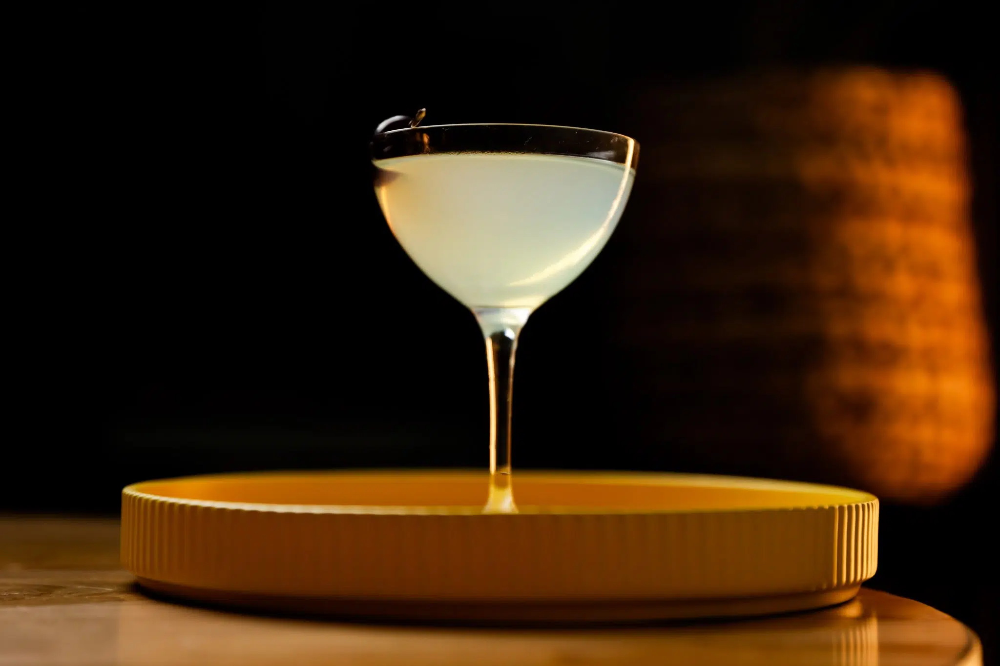 A side shot of a Closing Argument cocktail in a coupe glass on a yellow tray placed on a wooden surface