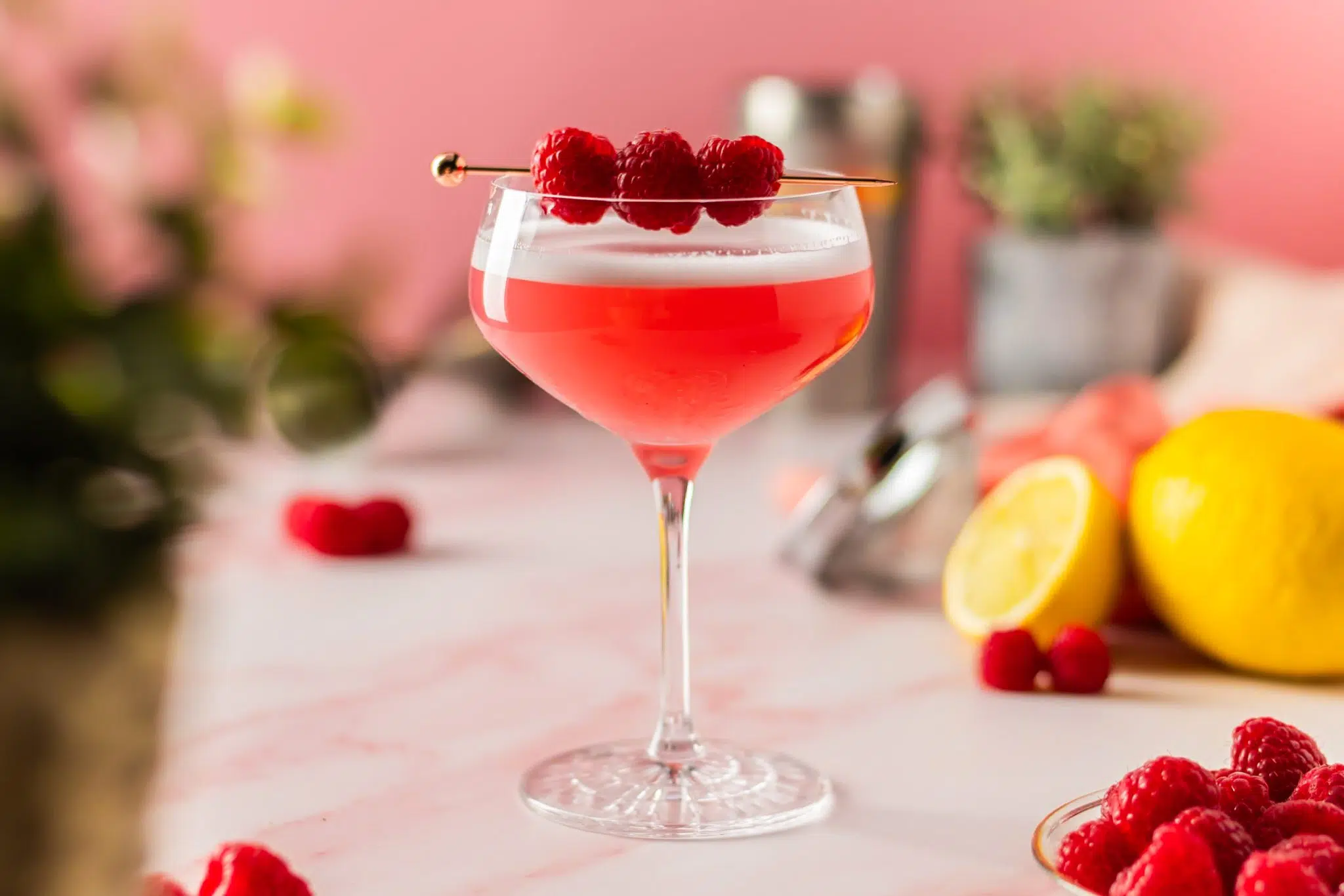 A side shot of a Cover Club cocktail in a cocktail glass on a white marmol table surrounded by fresh raspberries, two lemons, a plant and a shaker, in front of a pink wall.