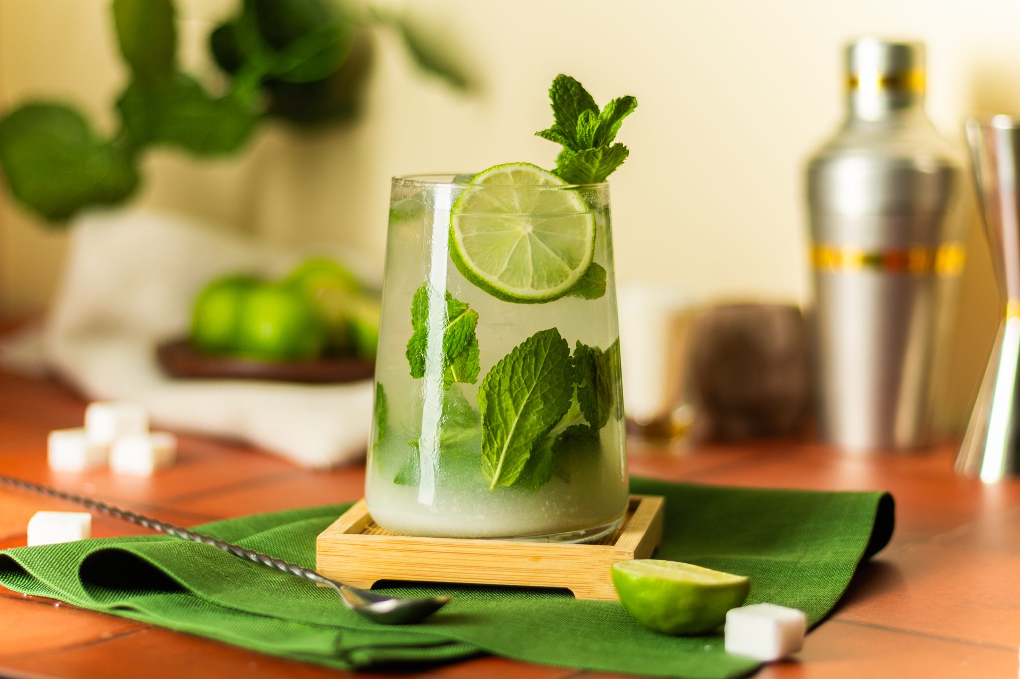 A side shot of a Coconut Mojito cocktail in a highball glass on a coaster placed on a green cloth on a tiled surface surrounded by sugar cubes, a bar spoon, limes, a shaker, and a white cloth, in front of a light yellow wall.