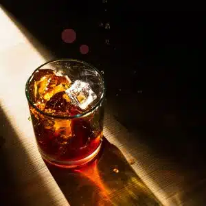 Old-fashioned glass filled with ice cubes and cognac illuminated by a ray of sunlight on a wooden surface.