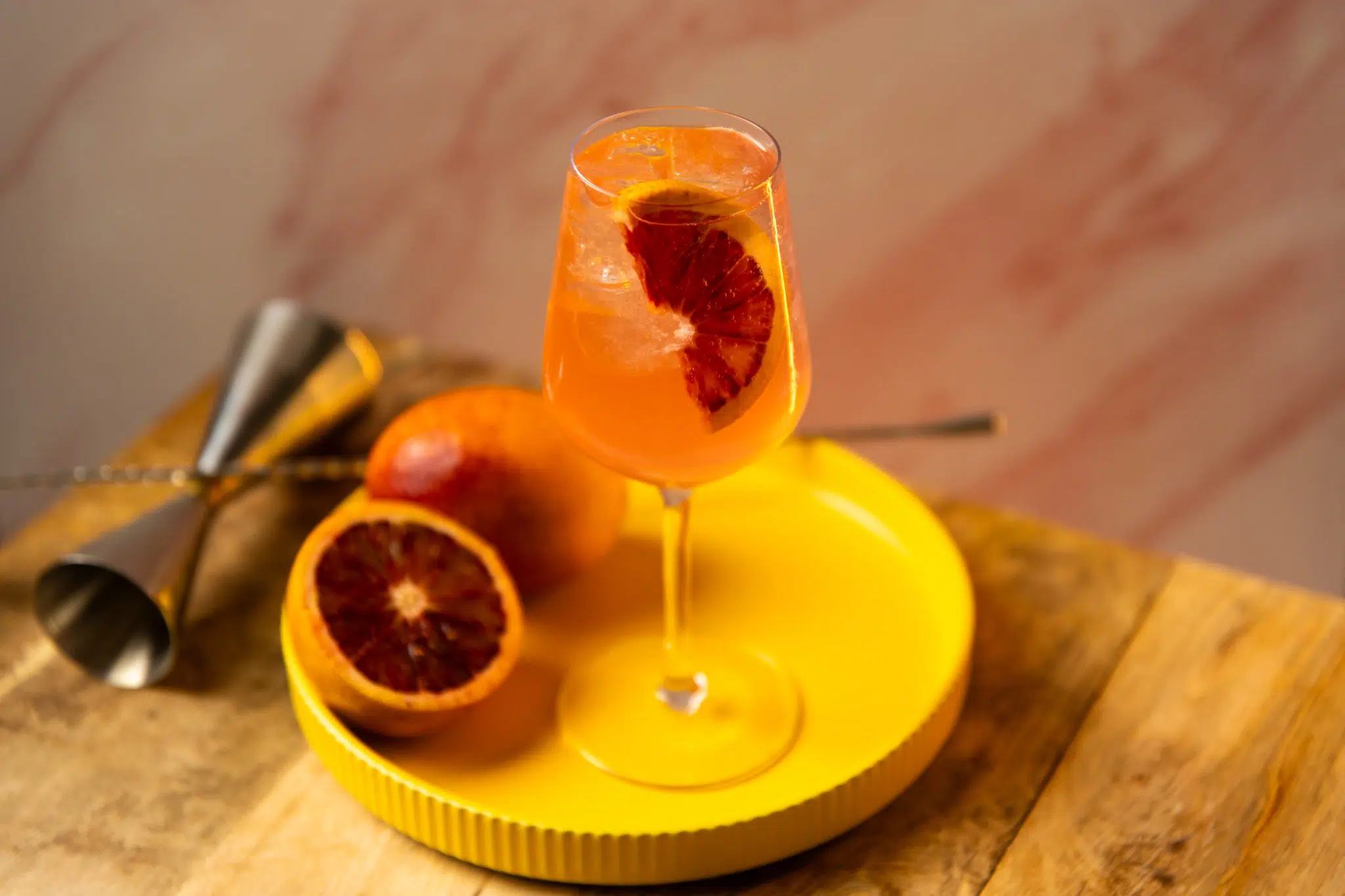 A side shot a Colletti Royale cocktail in a wine glass on a yellow tray with two blood oranges placed over a wooden table surrounded by a jigger and a bar spoon