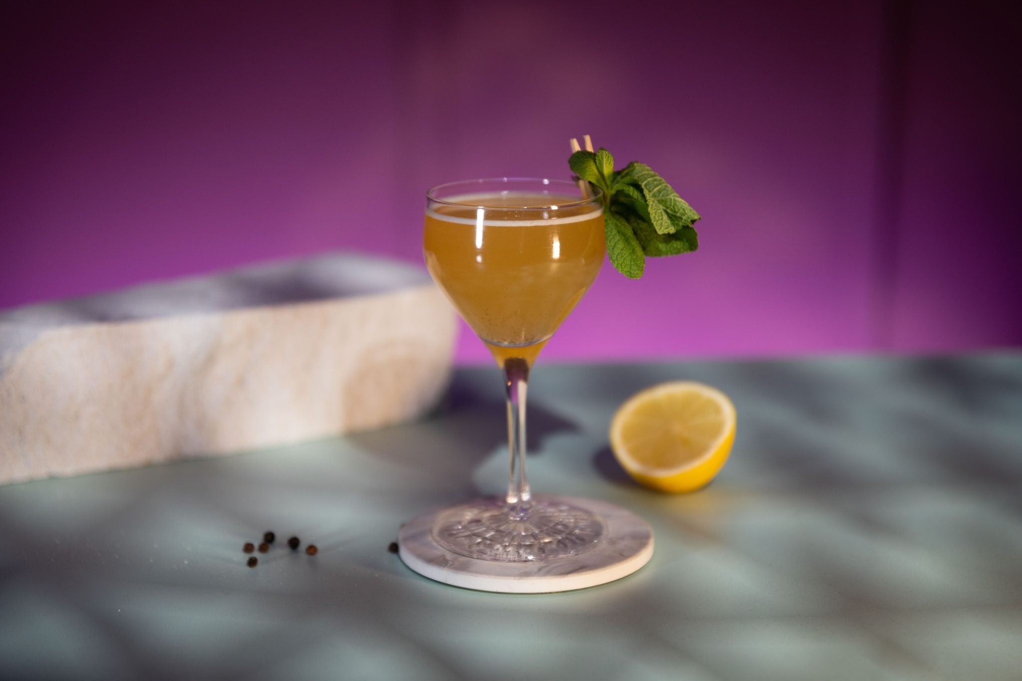 A side shot of a Companion cocktail in a Nick & Nora glass on a white coaster placed on a turquoise surface and a violet background surrounded by some black peppercorns and half a lemon