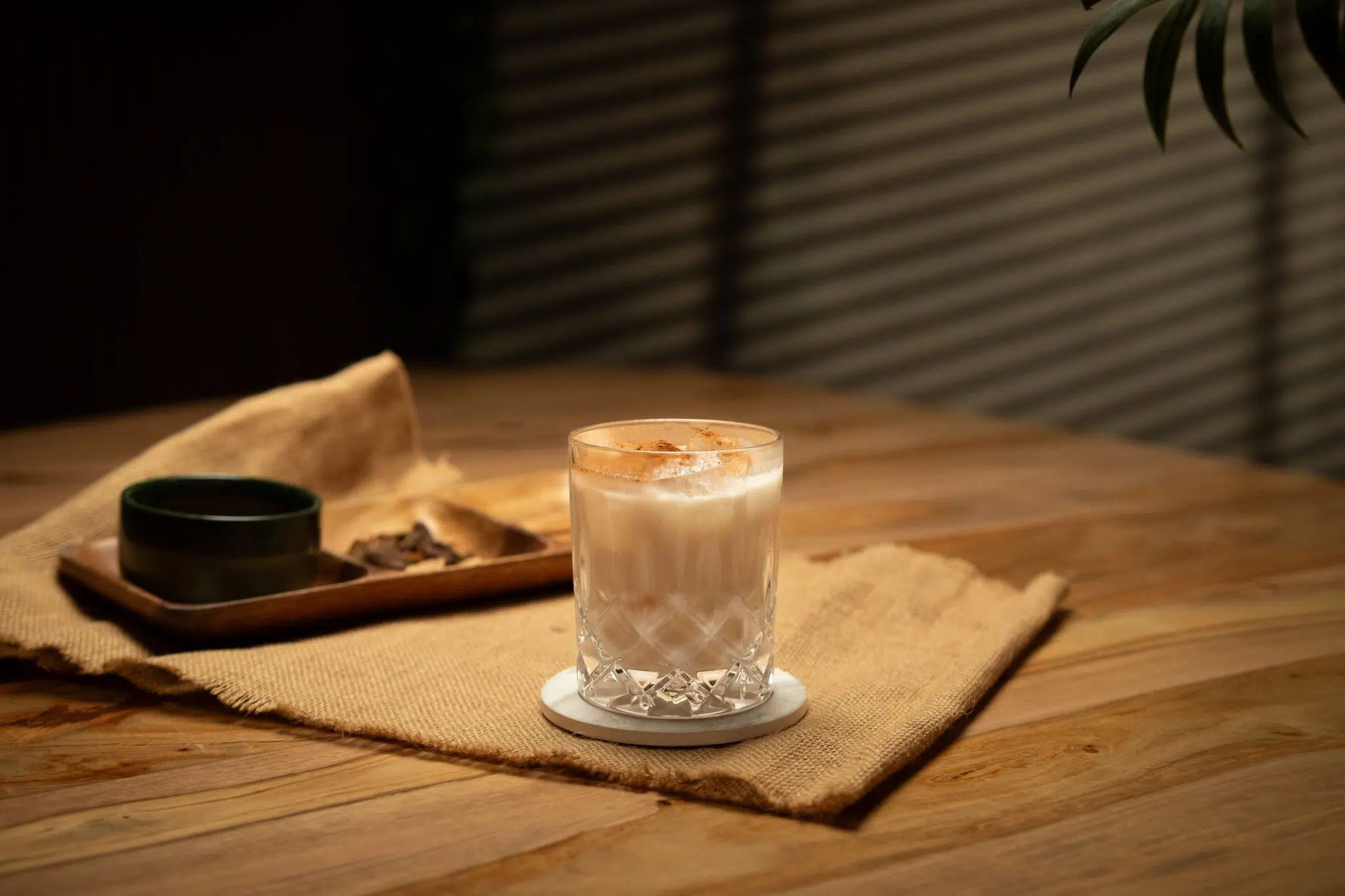 A side shot of a Coquito cocktail in a Rocks glass on a white coaster placed on a wooden table surrounded by a wooden tray with spices and a beige cotton placemat