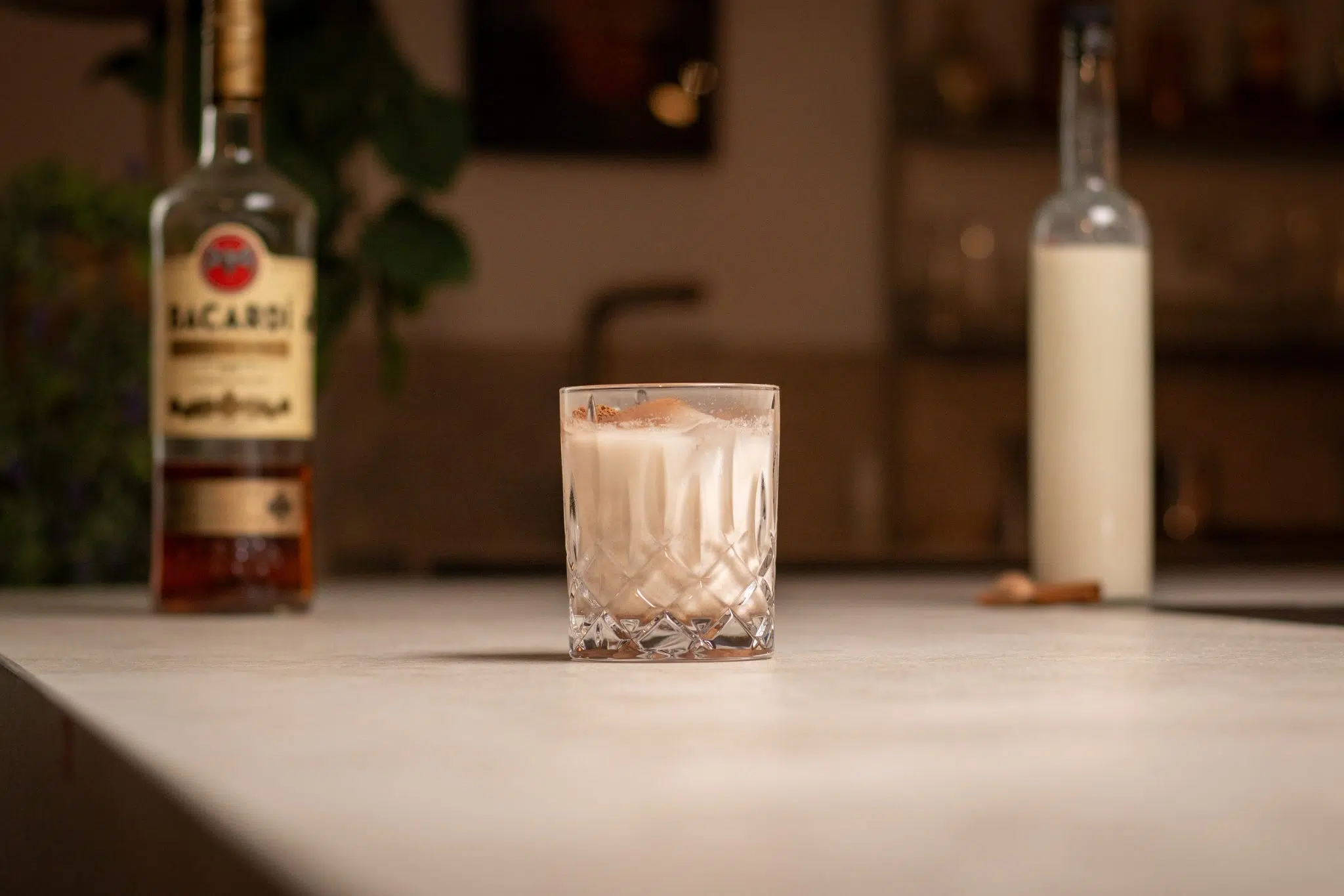 Dark Rum, Coquito mixture in a bottle, a cinnamon stick and a nutmeg laid out on a white bar table