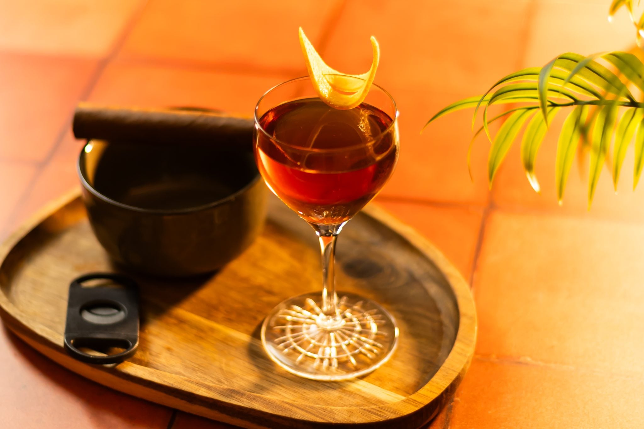 A side shot of a Corpse Reviver No.1 cocktail in a Nick&Nora glass on a wooden tray placed on a tiled surface with a black bowl with a ciger and a cigar cutter