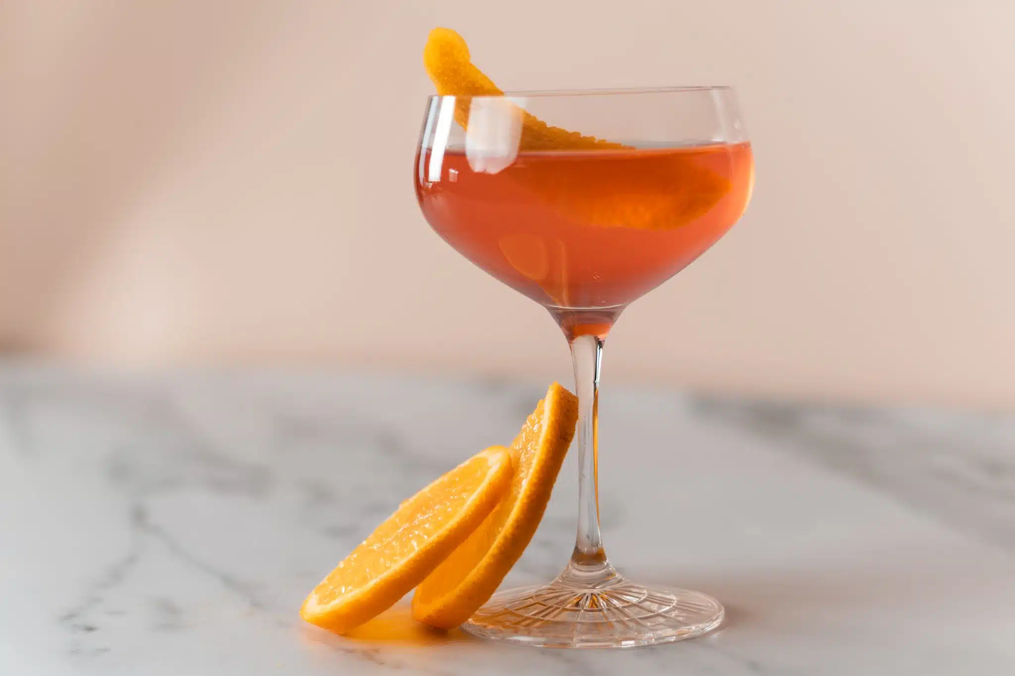A side shot of a Cosmopolitan cocktail in a martini glass on a white marmol table with two orange slices on the side.