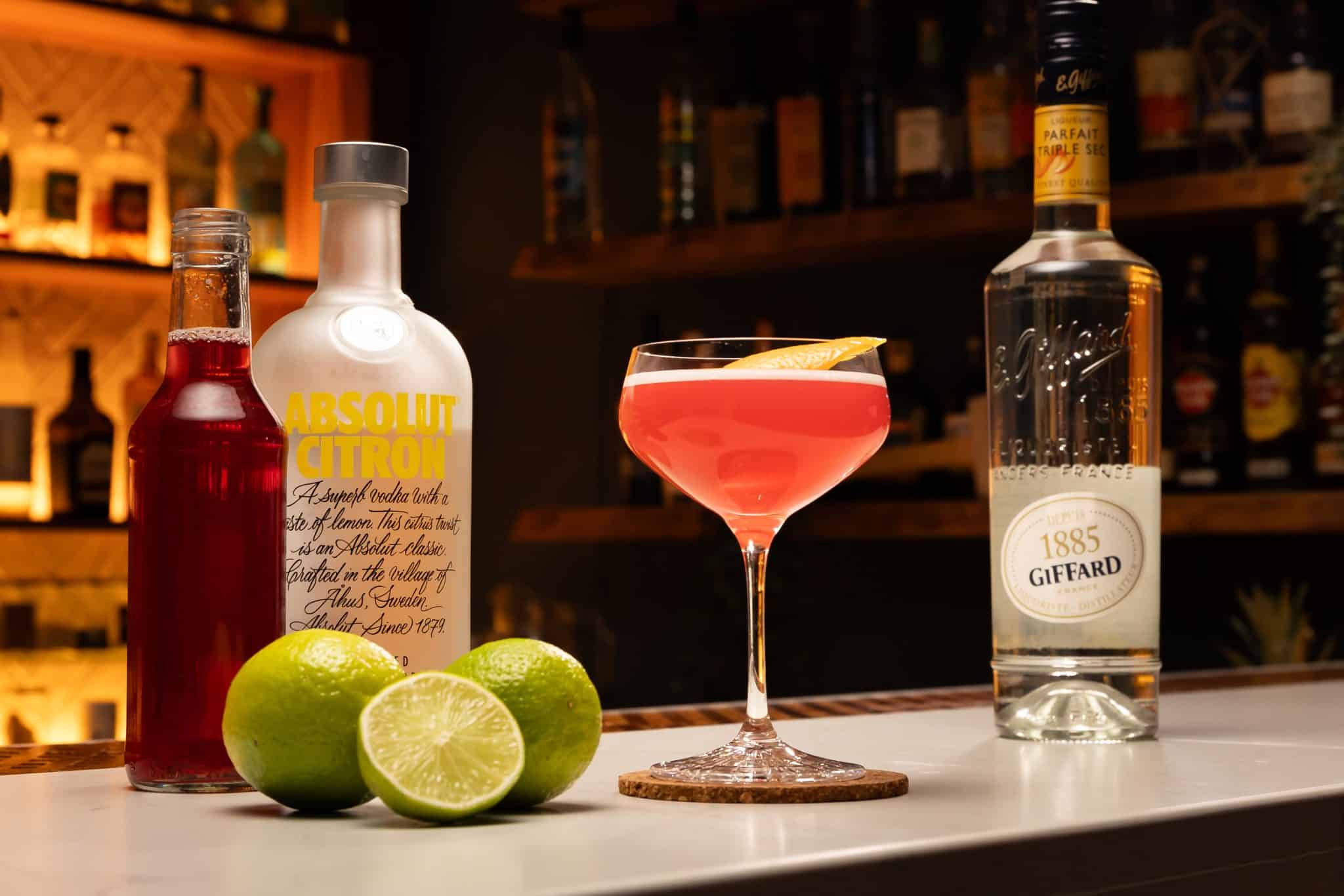 Vodka, triple sec, cranberry juice and lime juice laid out on a white bar table.