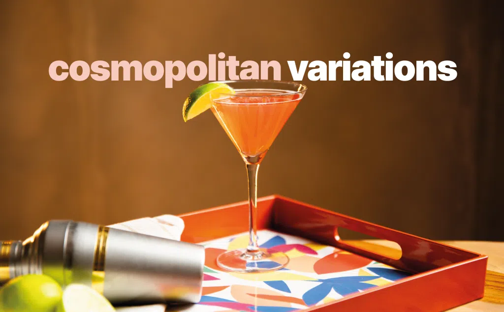 A side shot of a Cosmopolitan cocktail in a martini glass on a multicolor tray surrounded by a lime, and a cocktail shaker