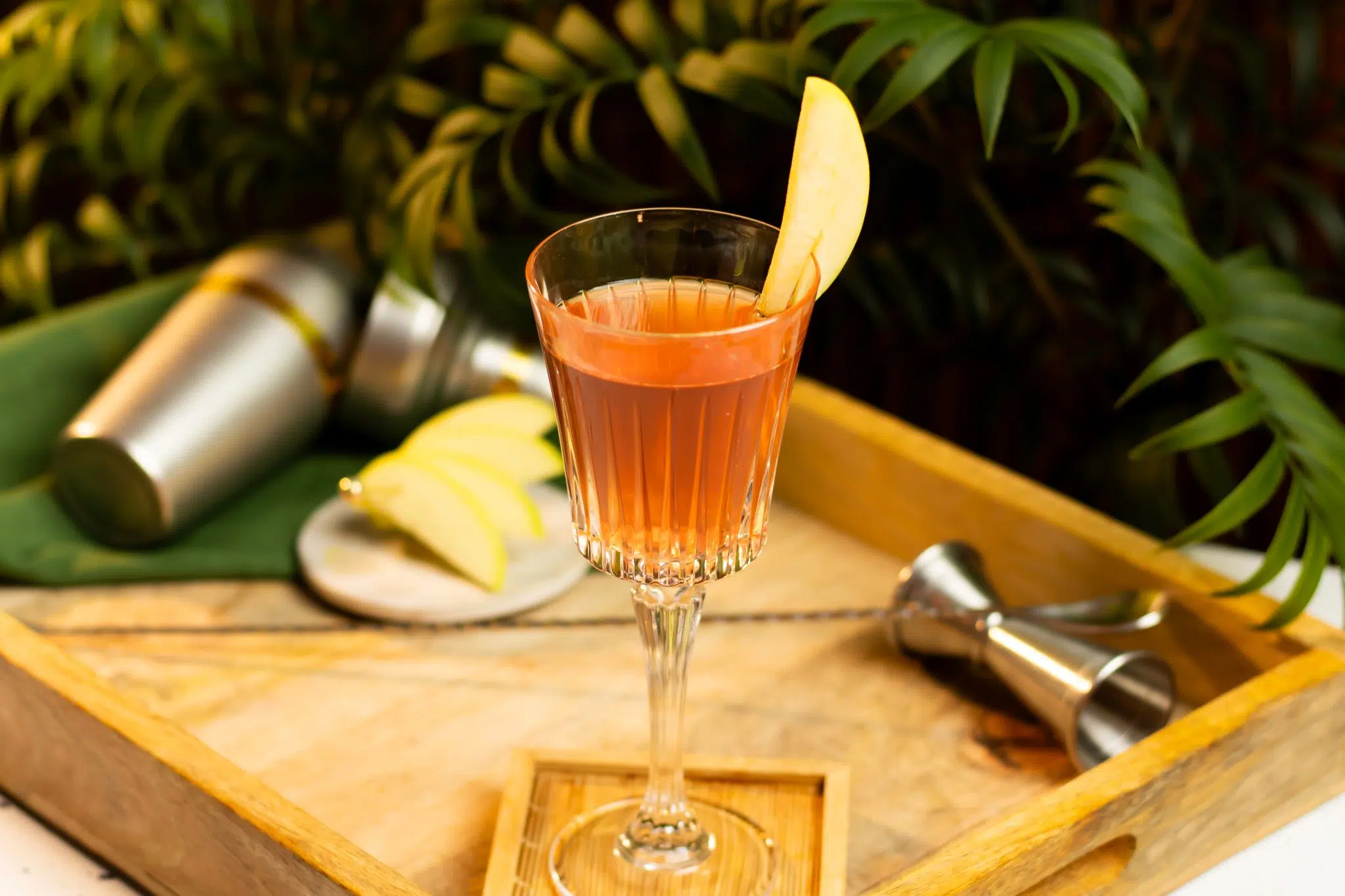 A side shot of a Cranberry Apple Mimosa in a champagne flute on a wooden coaster and tray surrounded by a shaker, a bar spoon, a jigger, a green cloth and a white coaster with apple slices