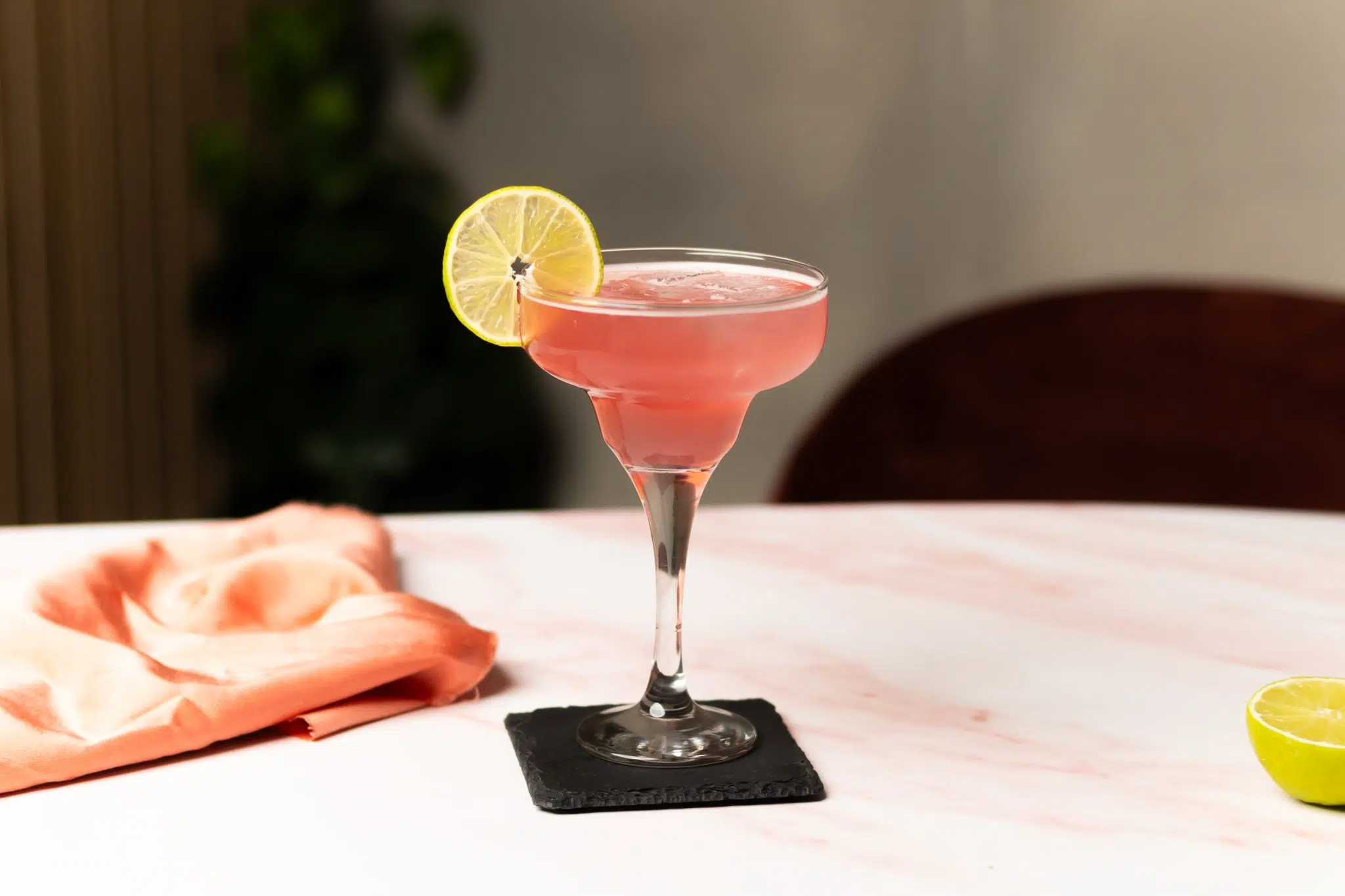 A side shot of a Cranberry Margarita cocktail in a margarita glass on a black stone coaster placed on a table surrounded by a salmon cloth and a lime.