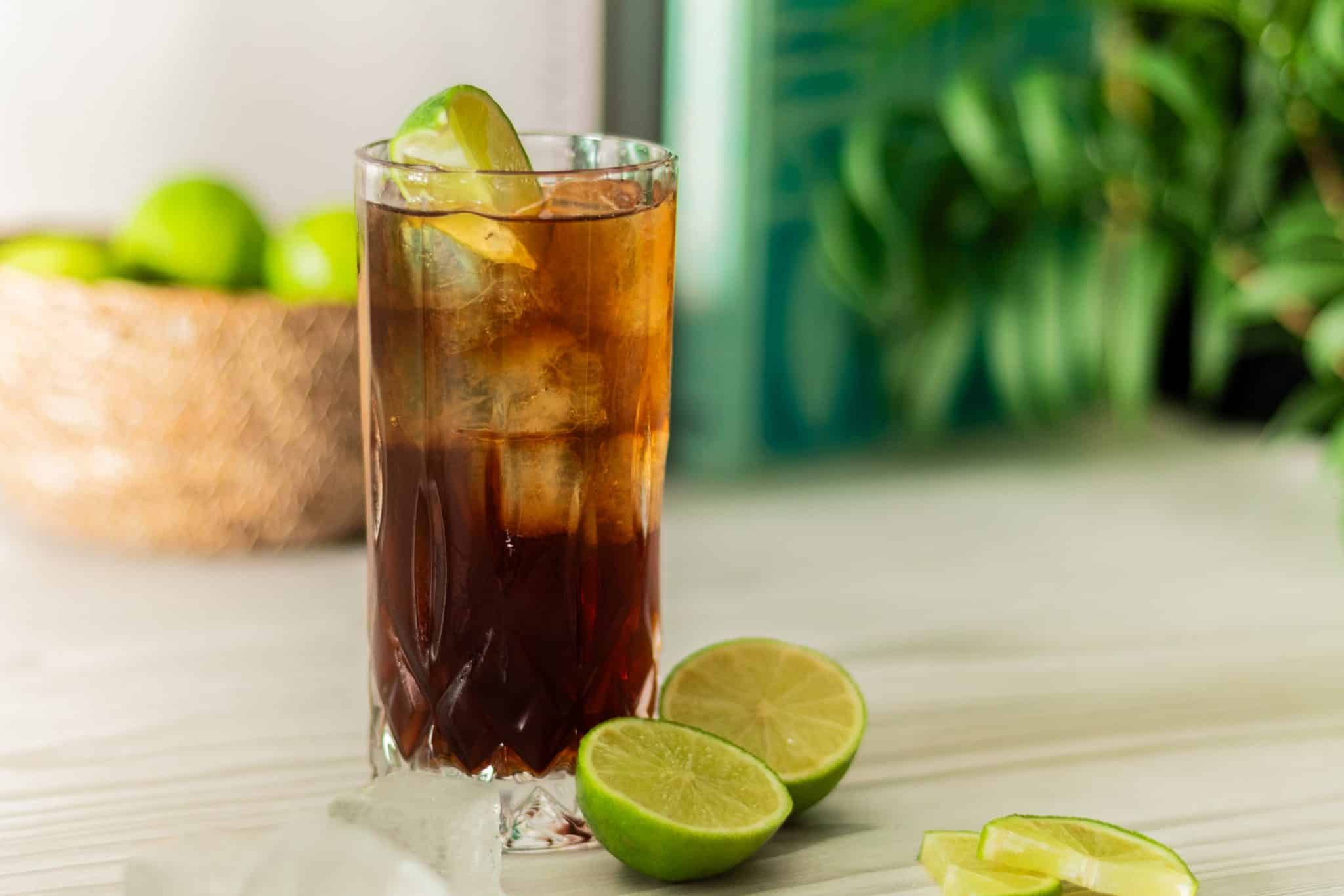A side shot of a Cuba Libre cocktail in a highball glass with two half limes on the side and a basket with limes on the background.