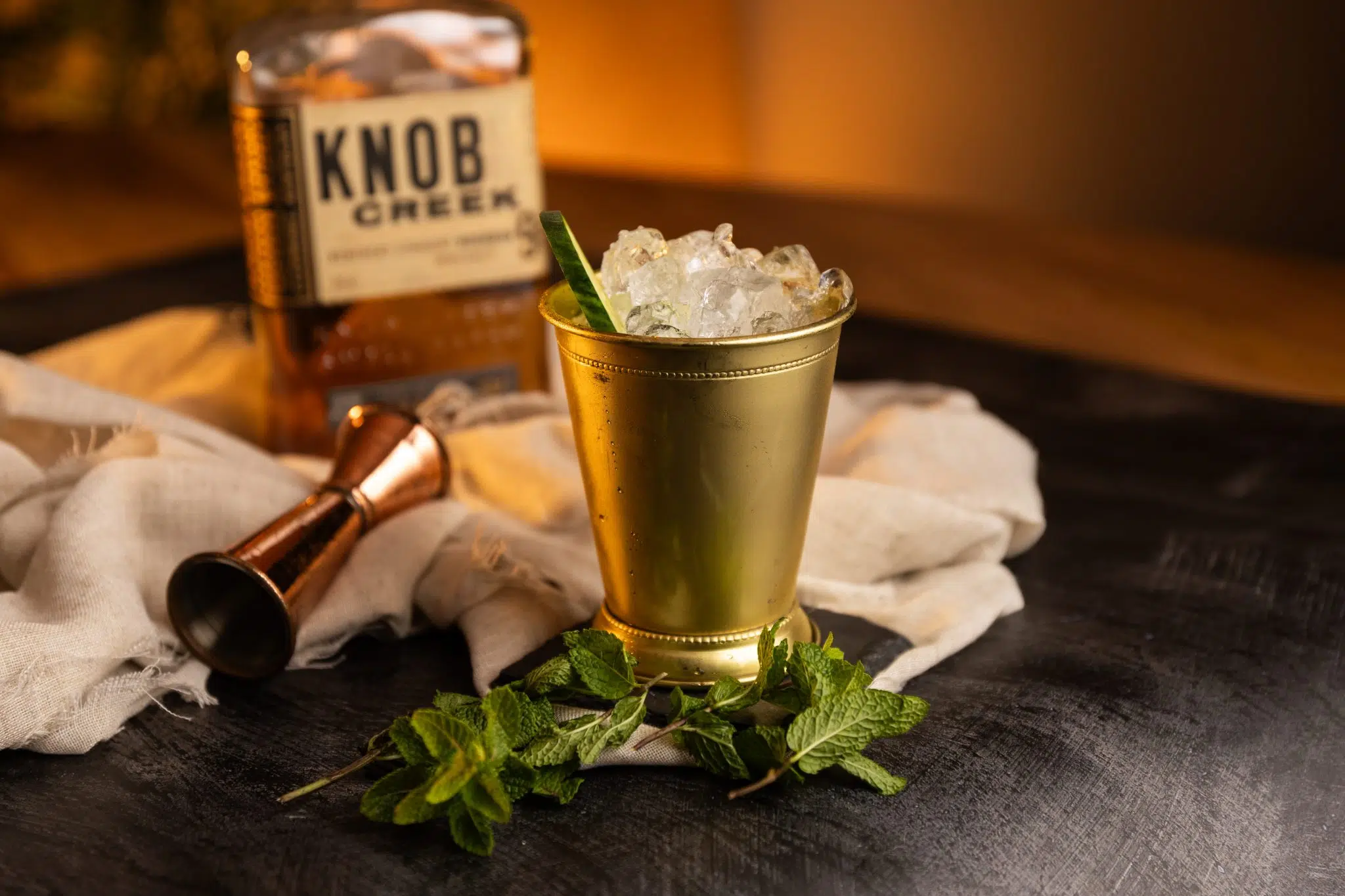 A side shot of a Cucumber Julep cocktail in a julep cup on a black stone coaster placed on a black surface surrounde by a white cloth, a jigger, mint sprig and a bourbon bottle