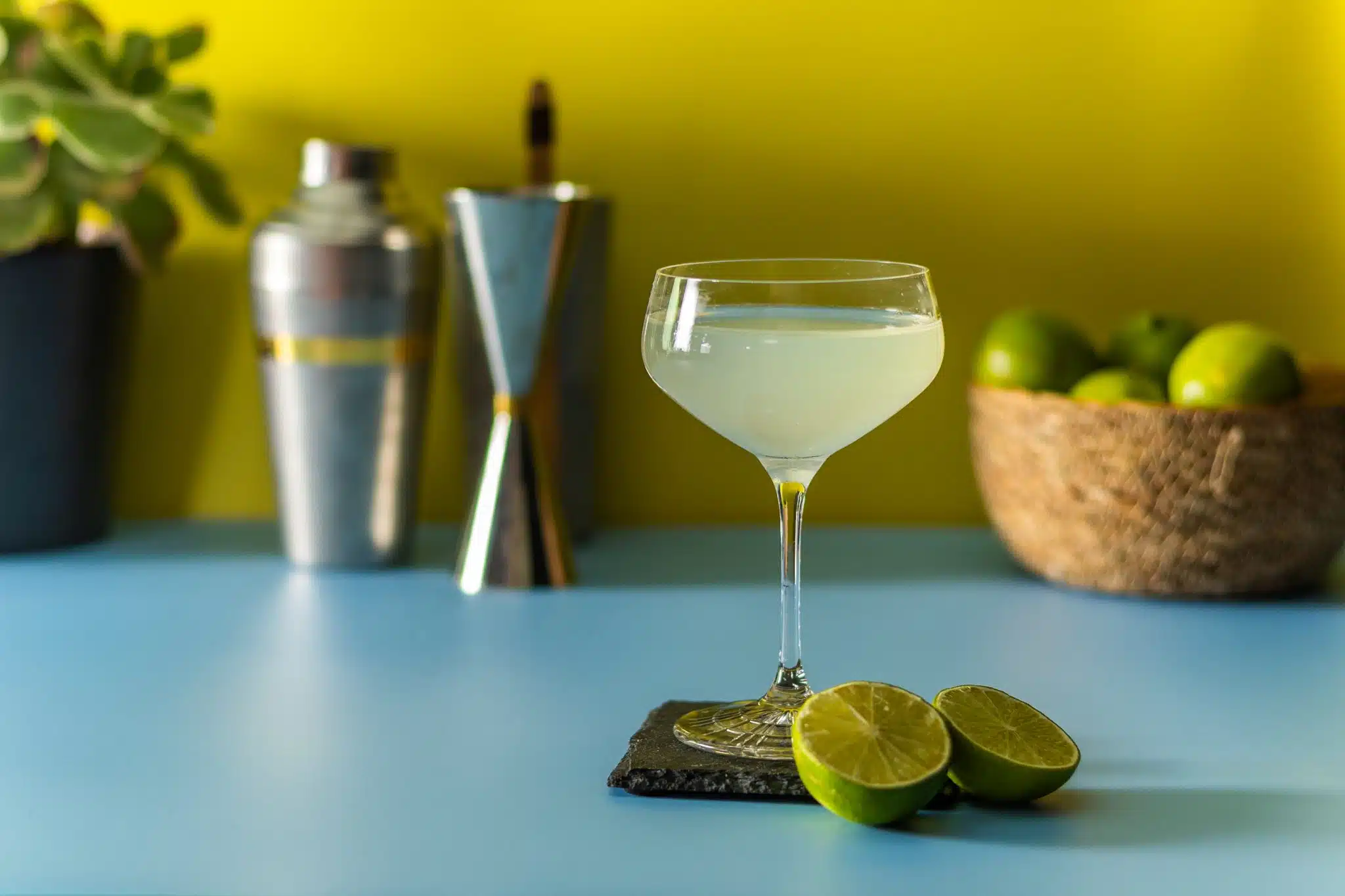 A side shot of a Daiquiri cocktail in a coupe glass on a black stone plate placed on a blue table with two half miles in front and a shaker, a jigger and a basket with limes on the background.
