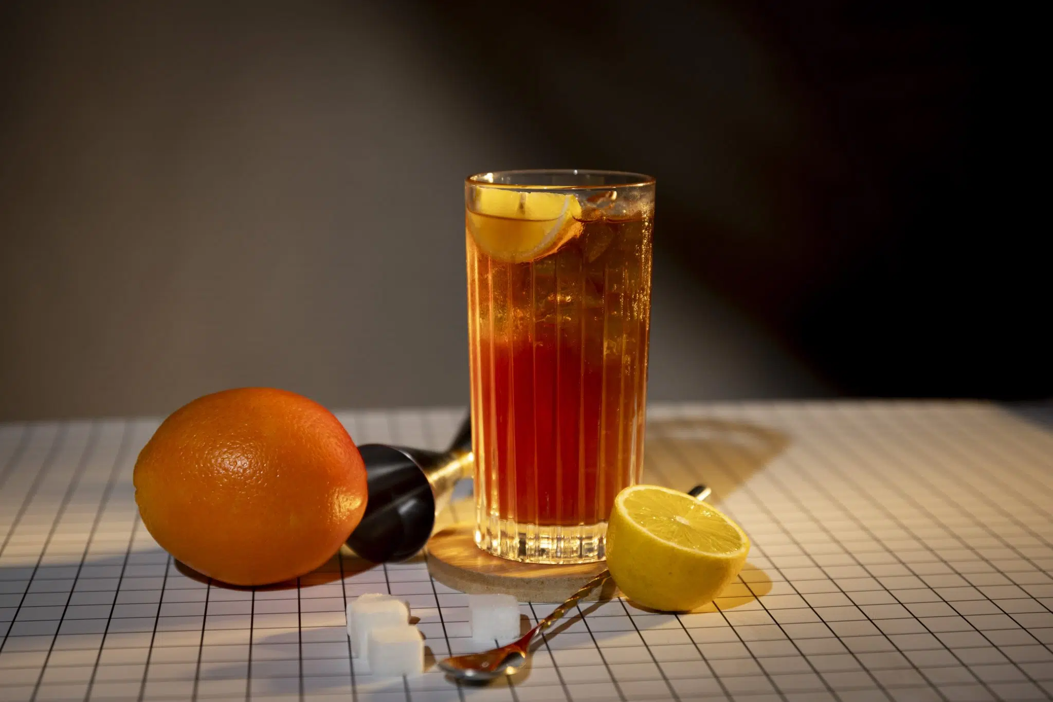A side shot of a Death Star cocktail in a highball glass on a wooden coaster placed on a checkered black and white tablecloth surrounded by an orange, half lemon, a jigger, three sugar cubes and a bar spoon