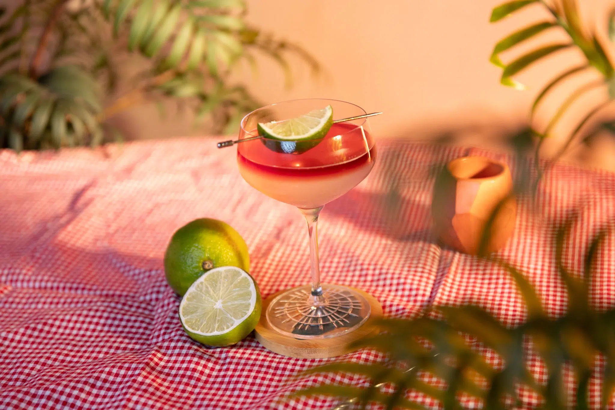 A side shot of a Devil's Margarita cocktail in a coupe glass on a wooden coaster placed on a red and white tablecloth surrounded by two limes and plant leaves